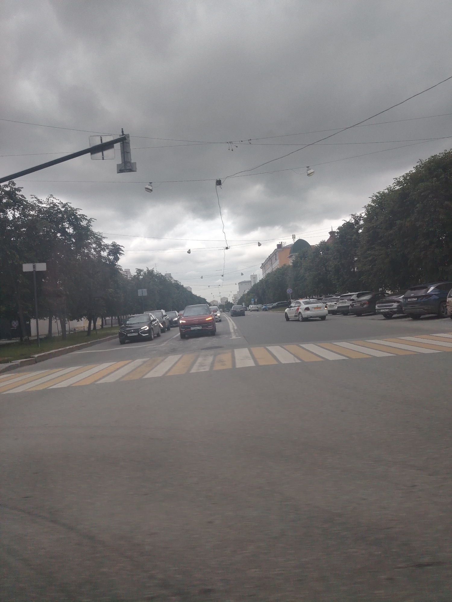 The image depicts a cloudy, overcast day in an urban environment. A wide asphalt road dominates the foreground, marked with a yellow and white striped pedestrian crossing. Several cars are visible on the road. In the middle ground, a line of cars is traveling away from the viewer, with a red SUV in the lead. To the left of the road, a sidewalk is lined with mature trees. A signpost is visible on the left side of the sidewalk. Across the road, on the right side, there is a row of parked cars and more trees. In the background, buildings of various heights and architectural styles are present. Overhead, power lines and traffic lights are strung across the street, with a single light fixture visible. The overall scene suggests a typical street view in a city on a cloudy day.
