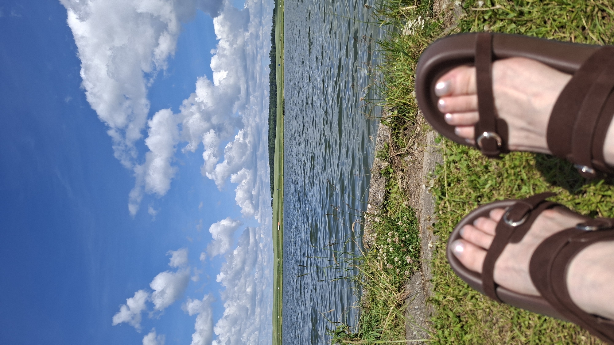 A first-person perspective shot shows a pair of brown sandals on a person's feet, which are resting on a grassy area next to a body of water. The person's toenails are painted with a pearlescent white polish. The water has gentle ripples and reflects the blue sky and scattered white clouds. In the distance, across the water, is a green, hilly landscape with a treeline and some small structures. The scene appears to be a serene outdoor setting, likely a park or recreational area. Based on the provided context, this location is in the city of Smaliavichy, Belarus.