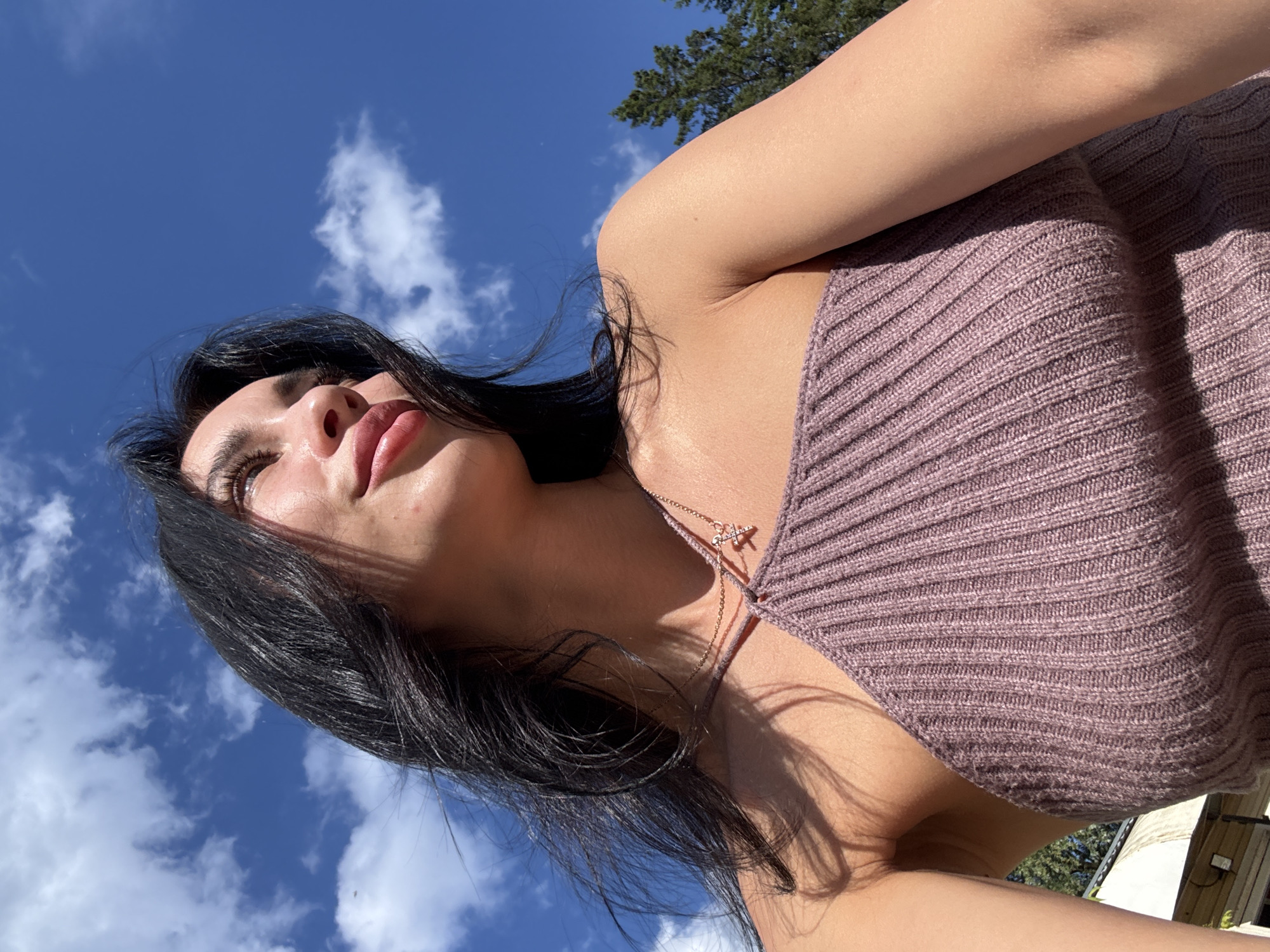 A young woman with dark hair and light skin is outdoors, looking up at the sky. She is wearing a mauve, ribbed knit top with a single strap. A delicate necklace with a small, star-shaped pendant is visible. The sky behind her is a vibrant blue with scattered white clouds. Sunlight casts shadows on her face and shoulder. The background includes the tops of evergreen trees and a glimpse of a building structure. The image appears to be taken from a low angle, emphasizing the sky. There is no discernible location in Chaikovsky, Russia.