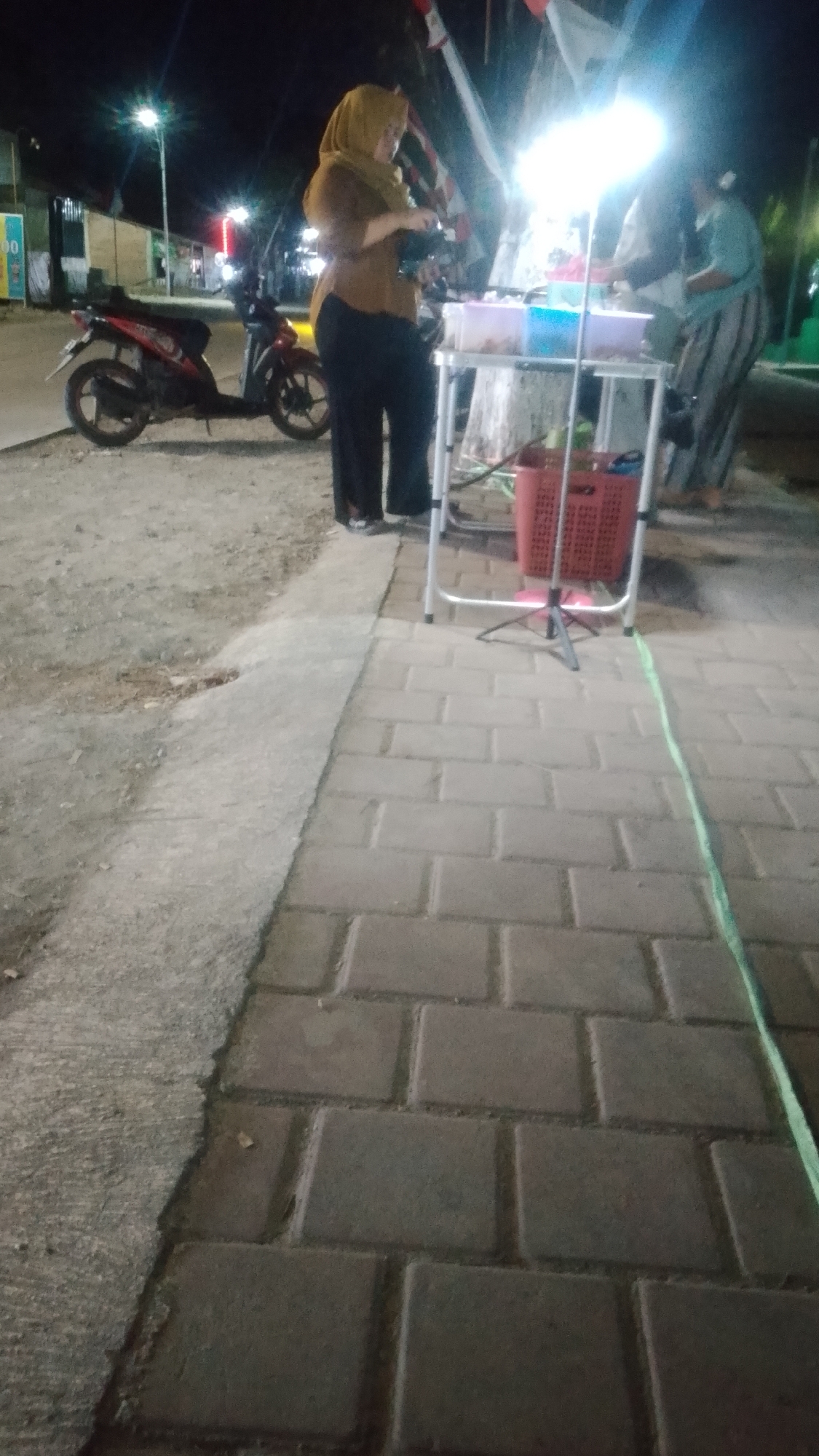 This is a nighttime photograph taken on a street in Karanglangu, Indonesia. In the foreground, a paved walkway made of grey rectangular bricks leads away from the viewer. To the left of the walkway, a dirt and gravel surface is visible.
In the middle ground, two women are standing near a portable food stall. The woman on the left is wearing a tan hijab and a dark brown long-sleeved shirt with black pants. She is holding a small item in her right hand and appears to be attending to the stall. The food stall itself is a metal-framed structure with a white tabletop. On the tabletop, there are several plastic containers filled with food items. A red woven basket is hanging from the side of the stall.
The woman on the right is wearing a light blue headscarf and a striped, loose-fitting garment with dark pants. She is also near the food stall.
Behind the food stall, a bright light illuminates the area. In the background, a red motorcycle is parked on the dirt surface. Further back, there are buildings and streetlights, some of which are illuminated. Red and white flags are visible, likely Indonesian flags, strung up and illuminated by the lights. The overall environment suggests a street-side food vendor operating at night.