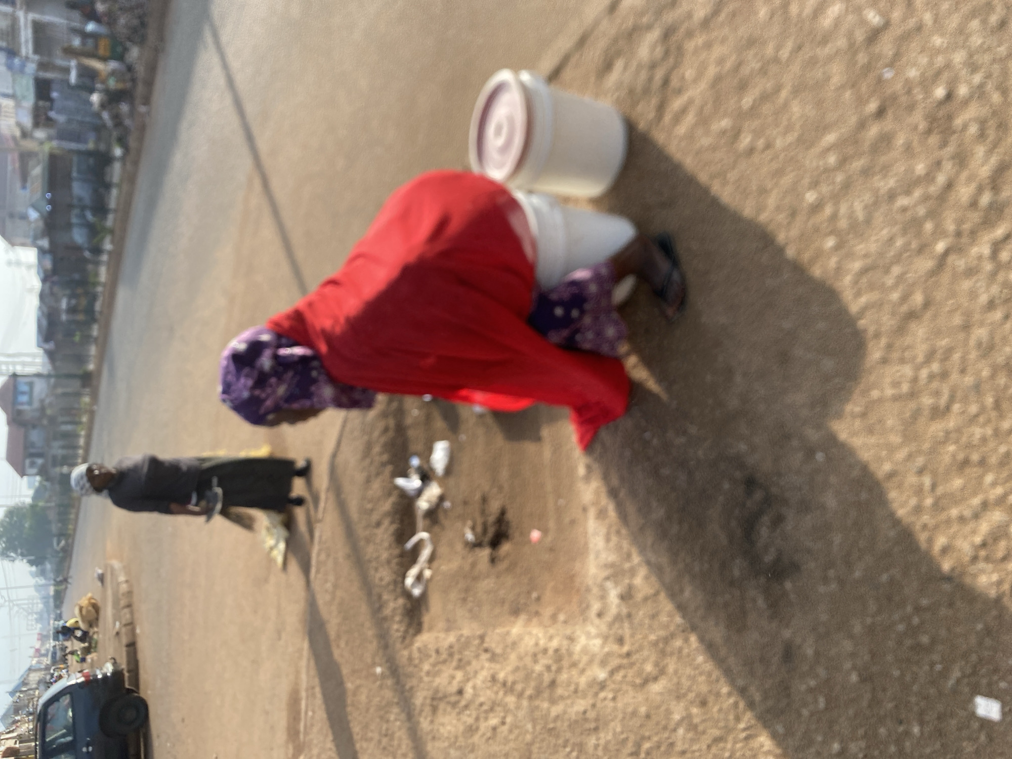 The image depicts an outdoor scene on a dusty street, possibly in Bukuru, Nigeria. In the foreground, a person wearing a red cloth and a patterned headscarf is crouching next to two white buckets, one of which has a reddish lid. The person's feet are clad in dark sandals. In the middle ground, another individual stands holding a broom and a piece of cloth. This person wears a dark top and a long, light-colored skirt, along with a headscarf. The ground is dirt and appears somewhat uneven, with some scattered litter. In the background, a dark-colored vehicle is parked on the left, and a row of buildings and shops lines the right side of the street. Several other figures and vehicles are visible further down the street, suggesting a busy market or commercial area. The lighting indicates it is either early morning or late afternoon, with long shadows cast on the ground.