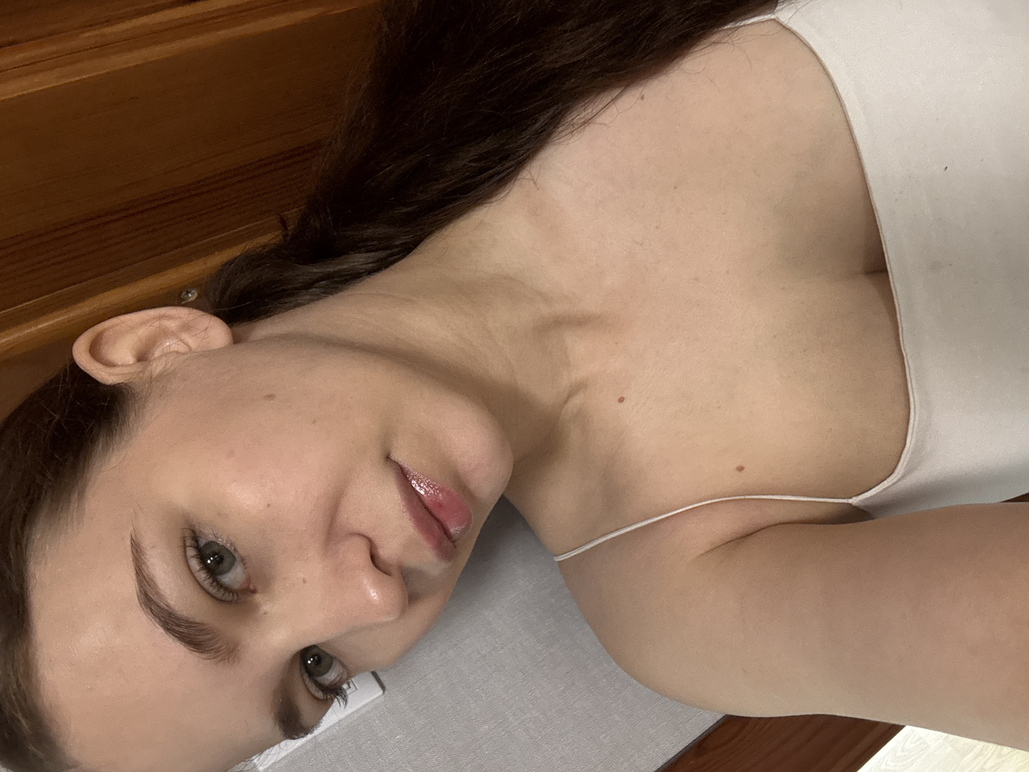 A young woman is depicted in a close-up photo, appearing to be lying down indoors and looking directly at the camera. Her head is tilted to her left. She has long, dark brown hair, light-colored eyes (appearing grey or green), well-defined eyebrows, and glossy lips with a soft, slightly parted expression. Her complexion is fair, with a few small moles visible on her neck and shoulder.
She is wearing a simple white camisole with thin straps. The background behind her head and upper torso features a dark brown wooden surface, possibly part of a dresser or headboard, with a small silver screw visible in the wood near her ear. She is resting on a light grey or white fabric surface, which could be a sheet or pillow.
The setting suggests a personal, relaxed indoor environment, likely a bedroom. The lighting is ambient, without specific indicators of the time of day. The overall mood is calm. No specific activities other than posing for the photograph are evident. No text is visible in the image. The provided location context is Chaikovsky, Russia.