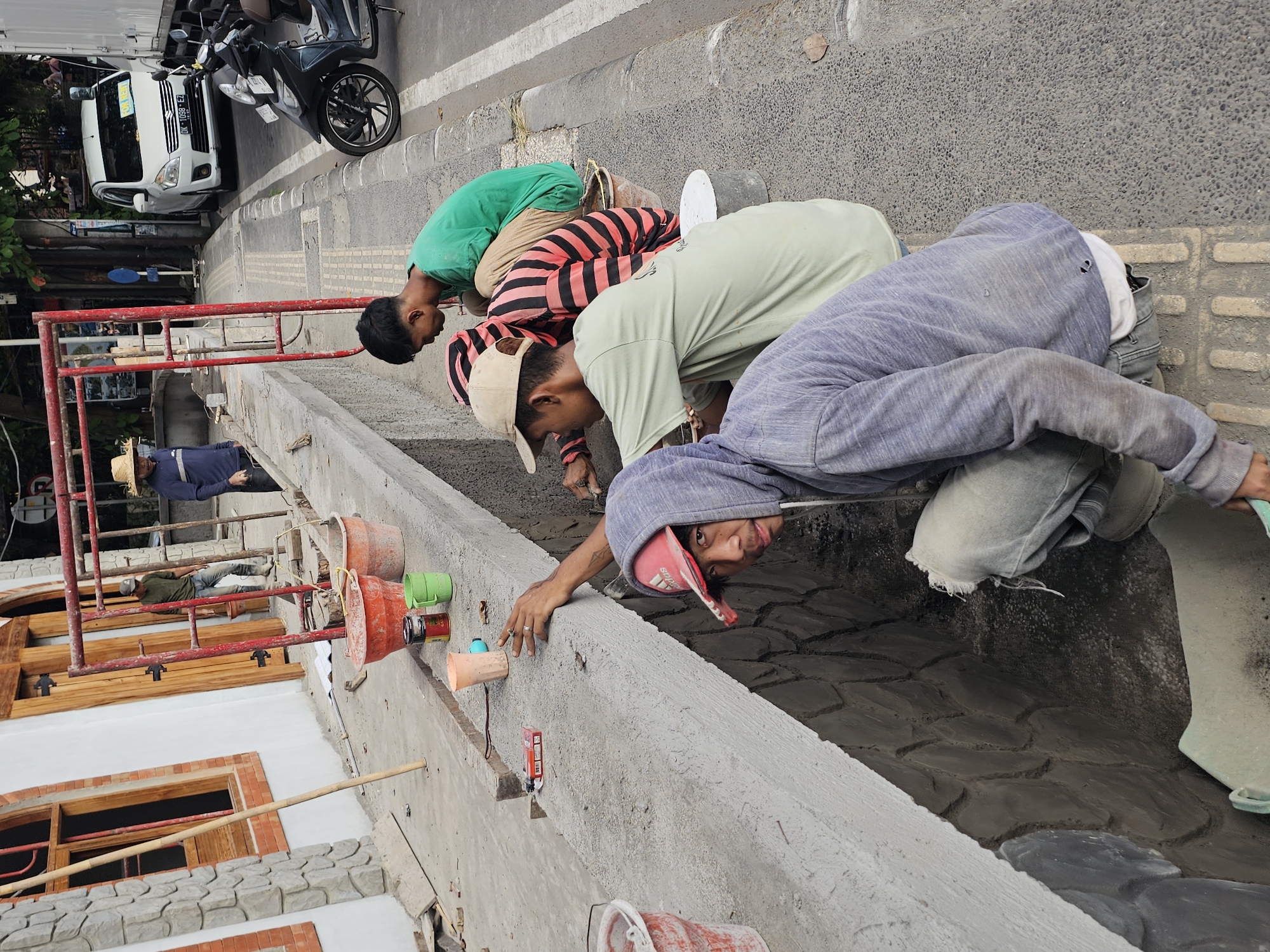 The image shows construction workers in Denpasar, Indonesia, working on a building facade. Four workers are visible, bending over a ledge, possibly applying mortar or finishing touches to a decorative wall treatment that mimics stone. One worker in the foreground is facing the camera. The facade of a building is partially visible, including window frames and red brick accents. Red metal scaffolding is present. Buckets and tools are scattered nearby. On the street, there are parked vehicles including a white van and a scooter. A person wearing a straw hat is seen lying on the scaffolding. The street is paved.