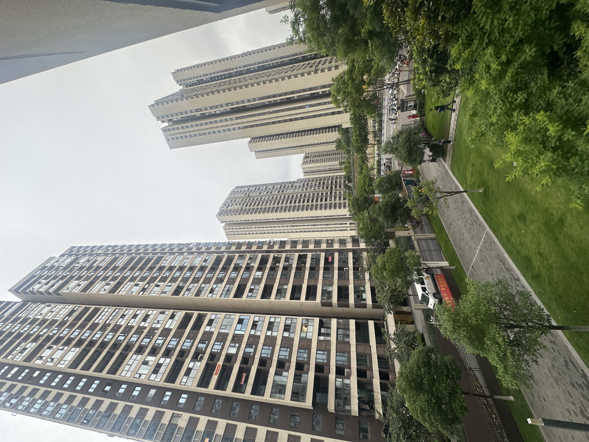 The image depicts an urban scene in Chongqing High-tech Zone, China, featuring high-rise apartment buildings and a landscaped public space. The buildings are modern, with repetitive window patterns. A paved walkway runs alongside a green lawn, interspersed with trees. People are present, walking in the distance. A vehicle is parked along the walkway. The sky is overcast.