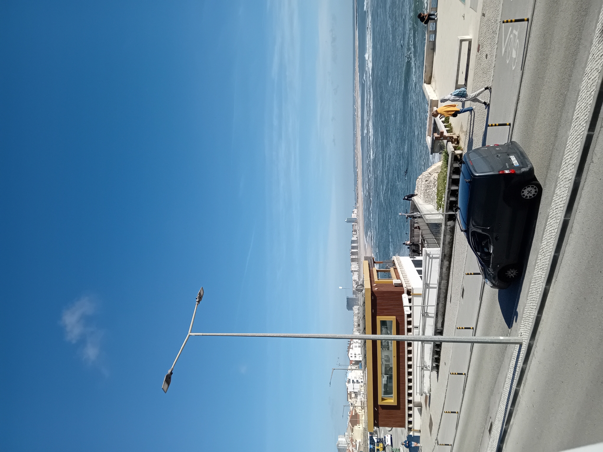 The image presents a street scene near a coastline under a clear blue sky. A black vehicle is parked adjacent to a pedestrian walkway with a bike lane marking. On the walkway, individuals are observed, one wearing a yellow top and interacting with the scenery while another sits on the edge looking out. Beyond the walkway is the ocean meeting a sandy beach, with city buildings visible in the distance. A lamppost is prominent in the foreground.