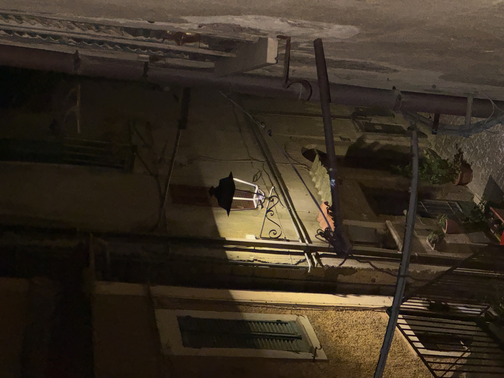 An exterior shot depicts a scene in Castelnuovo di Porto, Italy, at night. The focus is on the architecture of the buildings, including a mounted lamp and a window with shutters. The image is taken from a low angle, showing the underside of architectural structures, pipes, and conduits. Potted plants are also visible on the ledge of the building. The lighting is subdued, with the mounted lamp providing a strong point of illumination.