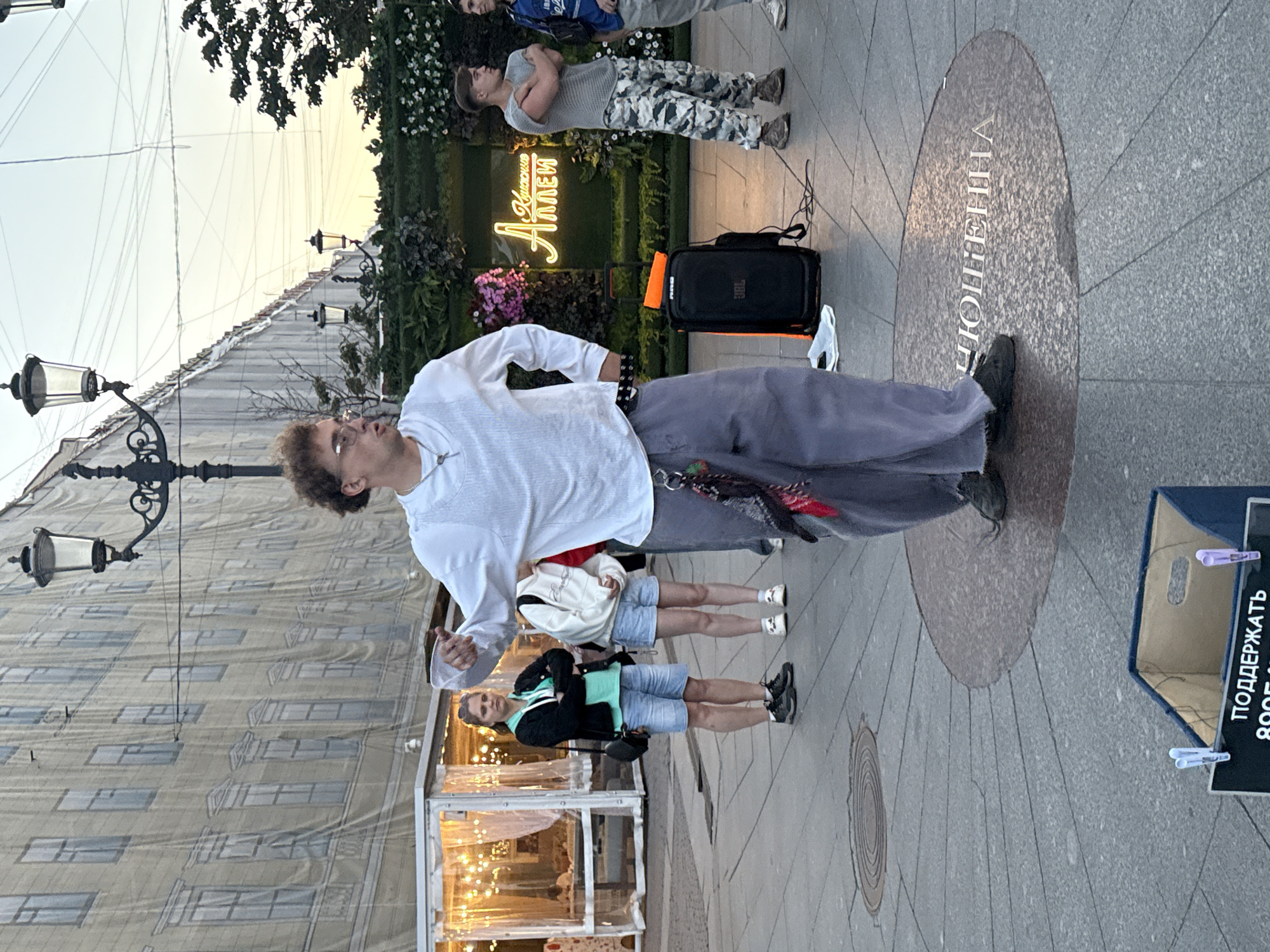 A medium shot shows a man in a white shirt and baggy gray pants performing on a paved public square in Saint Petersburg, Russia. The man, who appears to be a street performer, is mid-gesture, with his right arm extended and his left arm bent at the elbow. He has curly hair and is wearing glasses. In front of him is a black speaker with an orange item on top, and a blue box with text in Russian reading "ПОДДЕРЖАТЬ" (to support). Behind the performer, a woman in a black jacket and blue shorts stands with her arms crossed. Further back, a neon sign with Russian text that reads "Аллея" (alley) is visible, and two young men are standing near it. The background features a building draped with a large banner depicting windows, streetlights, and some greenery. The setting suggests a street or pedestrian area in a city, likely during daylight hours given the ambient light.