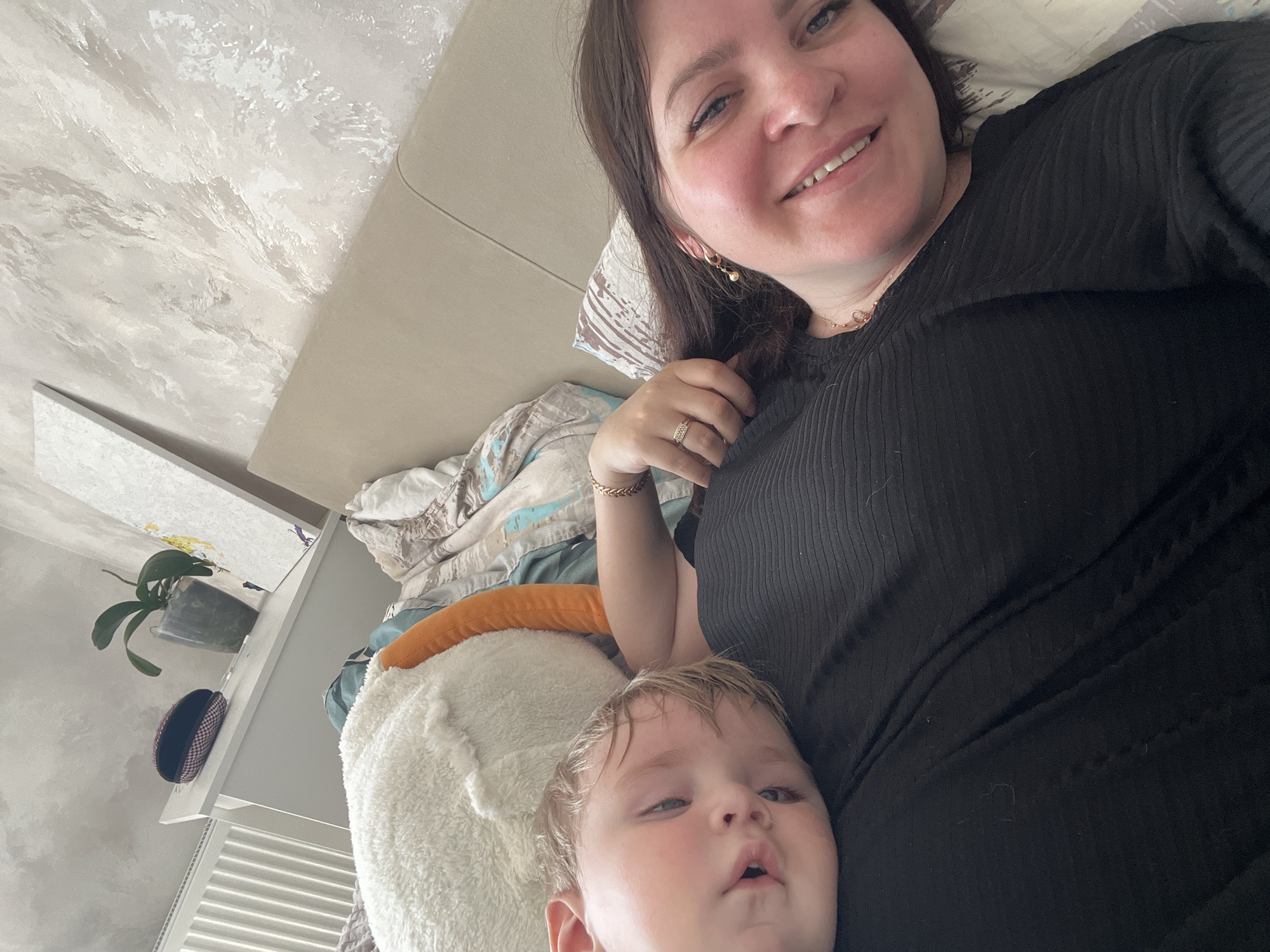 A woman and a toddler are lying on a bed. The woman is fair-skinned with brown hair and is wearing a black shirt. She is smiling and looking at the camera. The toddler is fair-skinned with blond hair and is looking at the camera with a neutral expression. The bed is covered with bedding and pillows. There is a nightstand next to the bed with a potted plant and a small box. The background is a wall with a textured pattern. The location is likely indoors, in a residential setting. There is no verifiable information to suggest that the image was taken in the city of Pervomaysky, Russia.