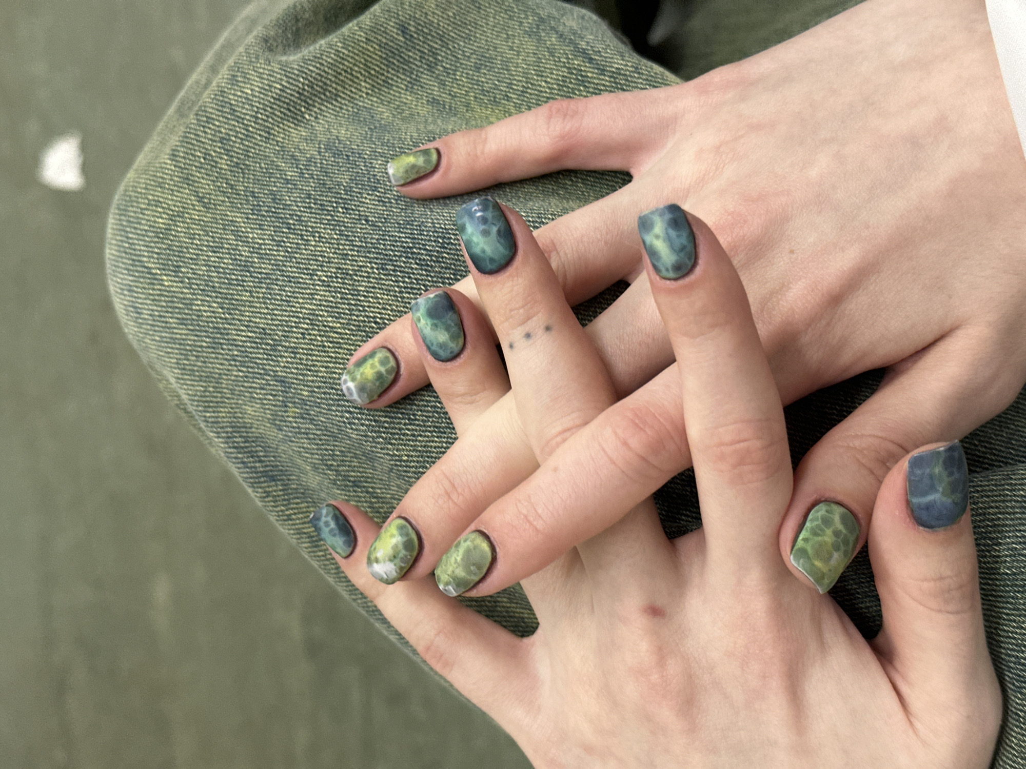A close-up shot features a pair of hands with artistic nail designs resting on what appears to be a lap covered in green denim fabric. The nails are painted in an abstract style, with colors ranging from blue to green and patterns that resemble organic textures, such as moss or geological formations. One hand shows three small dot tattoos between the knuckles. The scene's environment is nondescript, with the focus primarily on the hands and their decorative nails against the textile backdrop. There is no specific information provided indicating a location in Kommunarka, Russia.