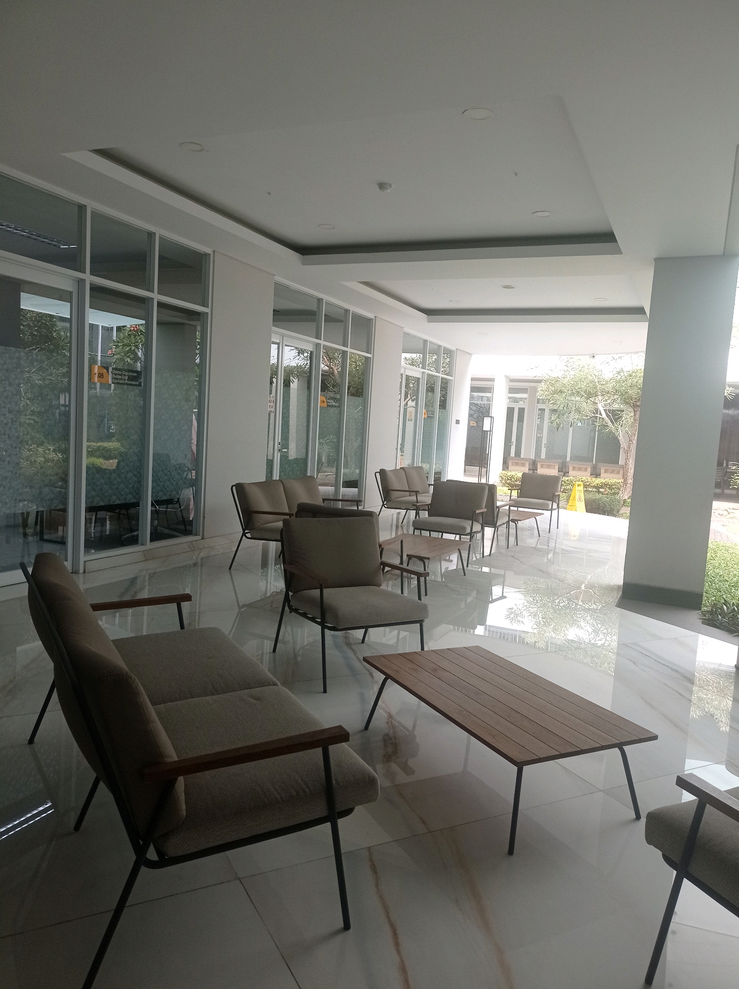 This is a full shot of an outdoor seating area in a modern building, possibly a waiting lounge or cafe, located in Bandung City, Indonesia. The area features a light-colored tiled floor with a glossy finish that reflects the surroundings. A covered walkway, indicated by the white ceiling and supporting pillars, extends into the distance. Large glass windows line the walkway, offering views of lush greenery and a tree outside.
Several seating arrangements are visible. In the foreground, a low, rectangular coffee table with a wooden top and dark metal legs is positioned centrally. To the left, a light-colored sofa with dark metal legs and wooden armrests faces towards the coffee table and the viewer. To the right of the coffee table, a single armchair of similar design to the sofa is partially visible.
Further into the seating area, more chairs and small tables are arranged. Several armchairs with light upholstery and dark metal frames are grouped together, some with small side tables. In the background, behind the seating area and beyond the glass windows, an outdoor space with more seating, planters, and a yellow caution sign is visible. The overall atmosphere is bright and airy, with natural light filtering into the space. The architecture is contemporary, with clean lines and a minimalist aesthetic.