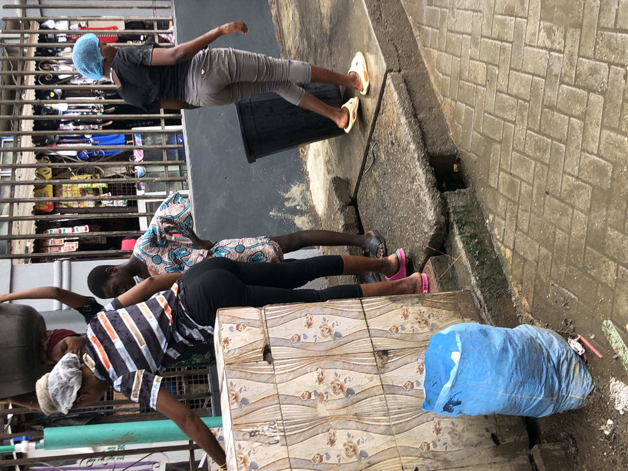 A group of people are gathered near a tiled wall and a metal fence with merchandise displayed behind it, possibly a market stall in Ikeja, Nigeria. In the foreground, a large blue trash bag is slumped on the wet ground. To the left, a woman wearing a striped shirt and black pants is leaning over, her face obscured by a hairnet and a hat. To her right, another woman in a patterned outfit is standing. Further to the right, a third woman, wearing a blue head covering, black t-shirt, and grey capri pants, walks away from the viewer. She is wearing light-colored clogs. The ground is paved with light-colored bricks and appears wet, suggesting recent rain. A large black trash bin is visible behind the woman in the blue head covering. The overall scene depicts a street or market environment with everyday activity.