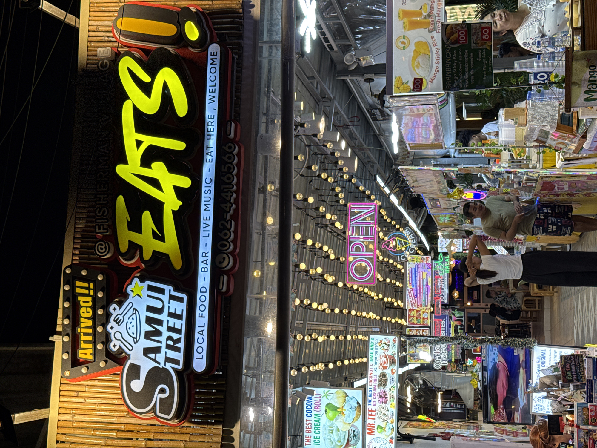 This is a nighttime photo of a vibrant street food market in Baan Bo Phut, Thailand. The scene is illuminated by numerous string lights and neon signs, creating a festive and lively atmosphere.
In the foreground, a large, illuminated sign for "SEATS!" stands prominently, with smaller signs advertising "Arrived!!" and "SAMUI STREET." The "SEATS!" sign also indicates "LOCAL FOOD - BAR - LIVE MUSIC - EAT HERE, WELCOME" and a phone number.
Further into the scene, various food stalls are visible, with signs advertising items like "Mango Sticky Rice," "Fresh Mango Juice," and "THE BEST COCONUT & THAILAND'S ICE CREAM ROLL." One sign specifically mentions "MR.TEE" ice cream. Neon signs with "OPEN" and other text in various languages are also present.
Several people are visible, including individuals working at the stalls and customers browsing or eating. One woman, seen in the upper right of the frame, is looking up and appears to be smiling. Others are partially obscured or in the background, engaged in their activities.
The overall impression is one of a bustling and inviting market, characteristic of a popular tourist destination in Thailand, likely during the evening or night given the lighting. The abundance of colorful signs and food offerings suggests a wide variety of culinary experiences available.