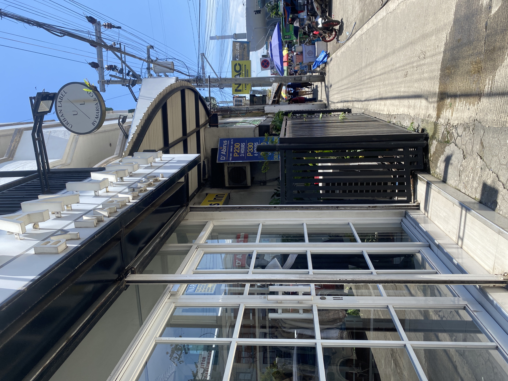 The image shows a street scene in Davao City, Philippines. In the foreground, there is a building with large glass doors and windows, likely a storefront. A black metal fence with vertical bars is visible to the right of the entrance. Above the entrance, a circular sign with the text "URBAN LABEL U L BY MARIA & CO." is mounted. Further down the street, there are power lines, utility poles, and other buildings. A sidewalk runs along the street, with several people walking and some street vendors with umbrellas and carts. Motorbikes are parked along the side of the road. The sky is clear and blue, indicating a sunny day. The overall impression is of a typical street scene in a commercial area of the city.