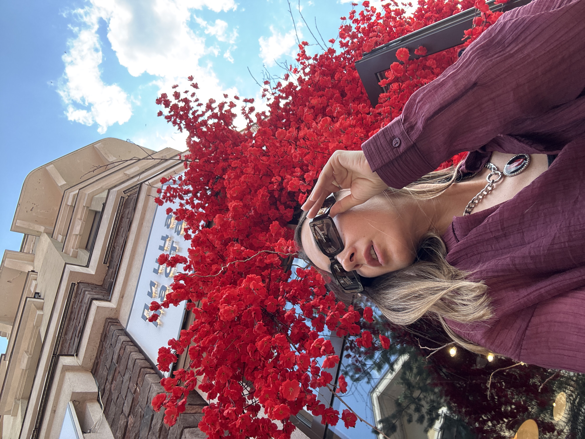 The image shows a low-angle, wide shot of a woman looking upwards and to her right. She is wearing a maroon, textured long-sleeved shirt and a chunky silver chain with a dark red pendant. She is also wearing dark sunglasses. Her blonde hair is visible, framing her face. The woman is positioned in front of a dense cluster of bright red decorative flowers, which are part of a tree-like structure.
Behind the flowers, a building with a light-colored facade and dark brick accents is visible. There is a white sign with dark lettering on the building. The sky above is bright blue with scattered white clouds. The overall lighting suggests it is daytime, likely sunny. The scene appears to be outdoors, possibly in an urban or commercial area.
The activity seems to be a casual moment of the woman taking a selfie or posing for a photo. The red flowers create a vibrant and dramatic backdrop. The location is identified as Estosadok, Russia. No specific event or emotion is clearly conveyed, beyond a general sense of enjoying the surroundings. There is visible text on the sign, but it is not legible enough to be transcribed.
