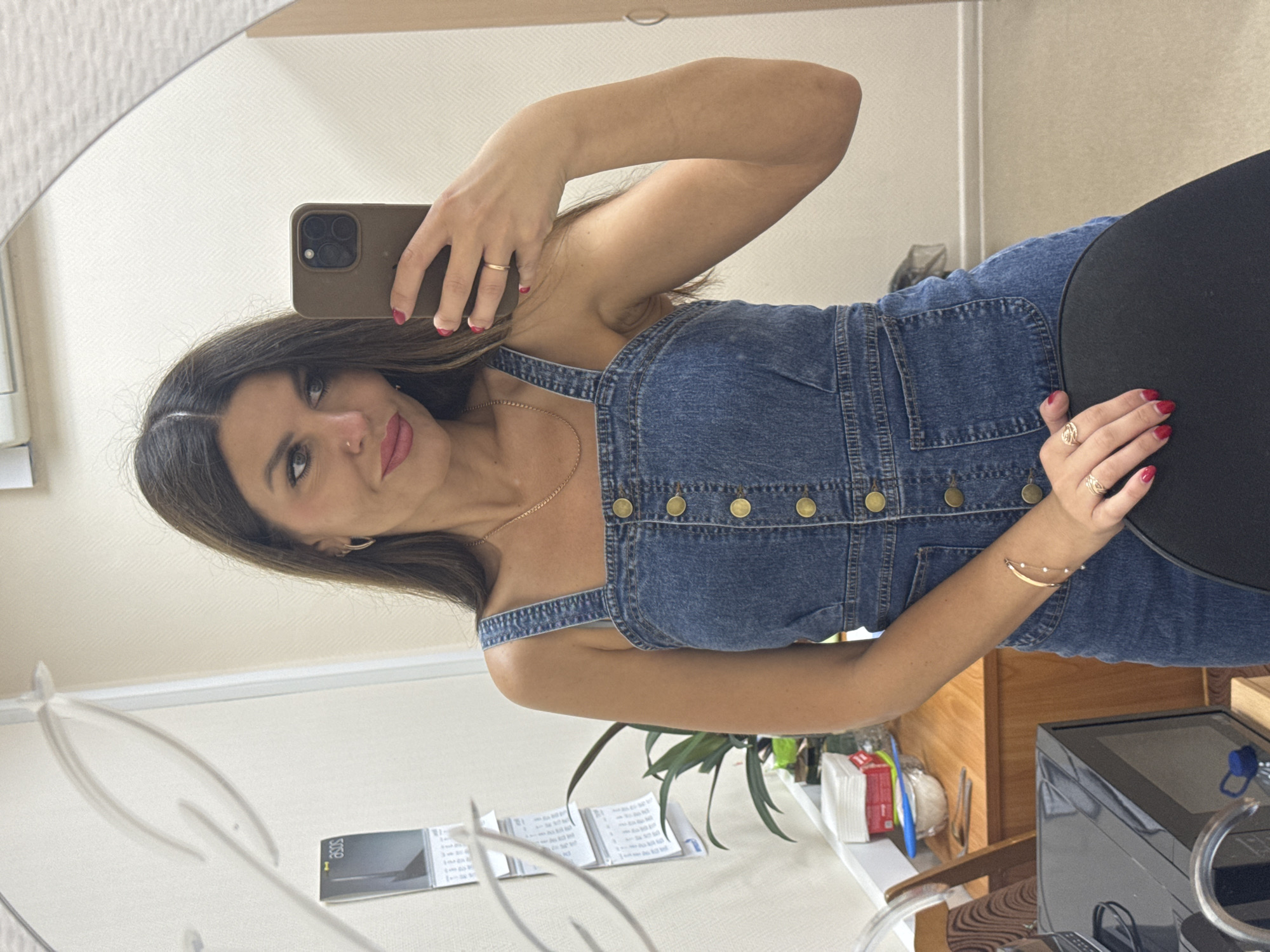A woman is taking a mirror selfie in what appears to be an office or domestic setting, potentially in Chaikovsky, Russia. She has long, dark brown hair parted in the middle, and is wearing a denim overalls dress with gold buttons. She is holding a smartphone in her right hand, angled to capture her reflection. Her left arm is resting on a black chair, and she is wearing a bracelet and rings on her fingers. In the background, a calendar is visible on the wall, along with a plant, a wooden cabinet with stacked papers and containers, and a microwave. The lighting suggests an indoor environment.