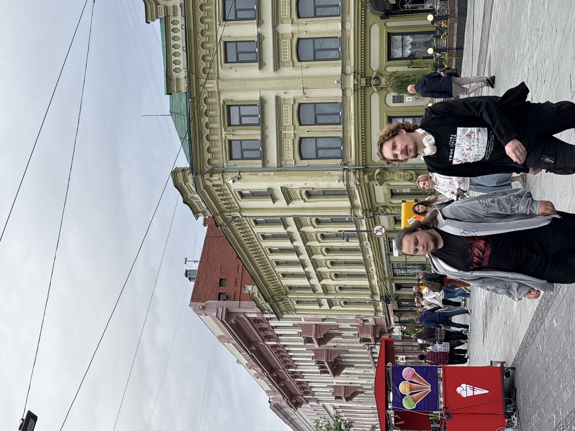 In this image, taken in Nizhny Novgorod, Russia, two young men are the primary subjects. One, with curly light brown hair and wearing headphones around his neck, is dressed in a black long-sleeved shirt with a graphic print and black pants. The other man, with longer brown hair, wears a black t-shirt with a red graphic and a grey hooded jacket over it, with black pants. They are walking on a paved pedestrian walkway.
The background features a row of multi-story buildings with ornate architectural details. The buildings have light-colored facades with numerous windows. A red ice cream cart with colorful graphics of ice cream cones and a white ice cream cone logo is visible in the foreground, suggesting the sale of ice cream. Several other people are walking in the background, indicating a public space.
The time of day appears to be daytime, as suggested by the ambient light, and the weather seems overcast. The overall scene depicts a casual, urban environment, possibly a city street or a public square, with people going about their day. No specific event is clearly indicated, but the presence of the ice cream cart and the general pedestrian traffic suggest a lively, public area. The expressions on the faces of the two main subjects are neutral, though the man with headphones appears to be looking towards the camera.