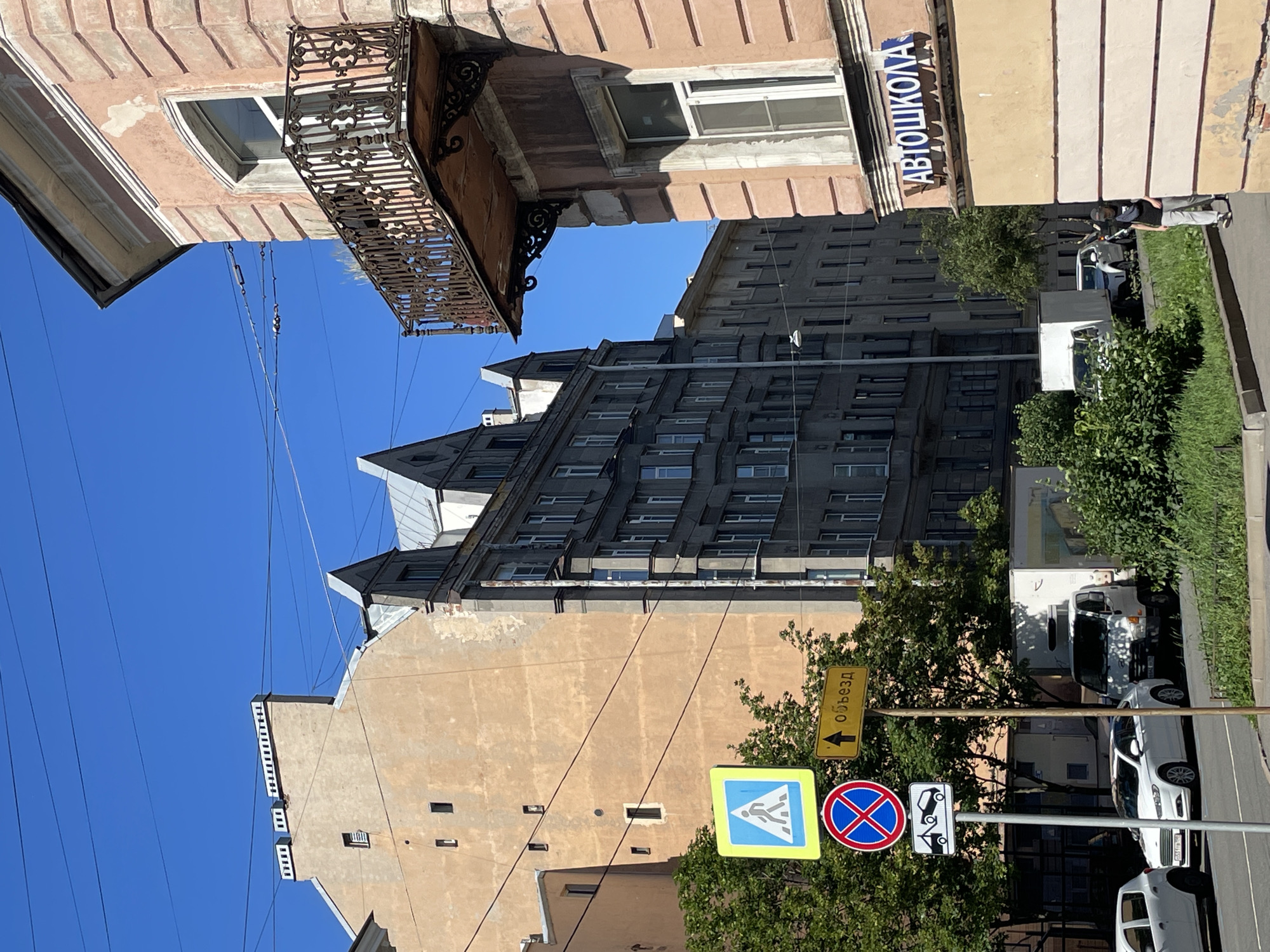 This is a street scene in Saint Petersburg, Russia, featuring a clear blue sky and multiple buildings. In the foreground, on the right, is a light pink building with an ornate balcony and a window. A blue sign with white Cyrillic text, "АВТОШКОЛА" (Autoschool), is mounted on this building. To the left of the pink building, a darker, tall apartment building with many windows is visible. Further to the left, a lighter colored building with a rough facade is seen.
Several traffic signs are present on metal poles: a blue square sign with a white pedestrian silhouette indicating a crosswalk, a circular red and blue sign with a red cross, and a rectangular sign below it showing a tow truck. A yellow rectangular sign with a black arrow pointing upwards and the word "объезд" (detour) is also visible.
Several cars, including white sedans and a white delivery truck, are parked or driving on the street. Green trees and bushes line the side of the road. A person is walking on the sidewalk in the lower right portion of the image, wearing a dark cap, dark scarf, gray t-shirt, and light-colored pants. Overhead, power lines are strung across the sky. The scene appears to be captured on a sunny day.