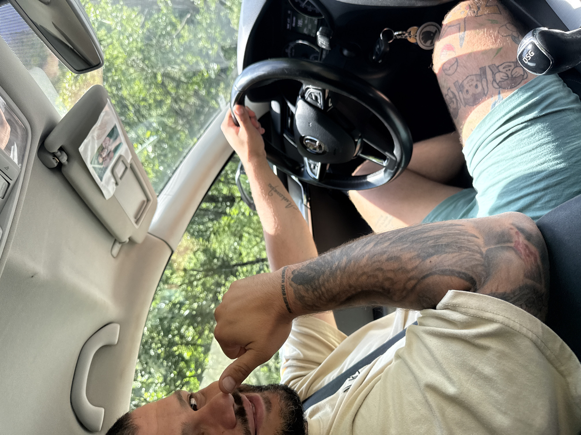 This image captures a low-angle, sideways view inside a car in Dorkovo, Bulgaria, during what appears to be a sunny day. A man is in the driver's seat, his face visible with dark hair and a beard, looking towards the camera. He is wearing a light-colored t-shirt and green shorts.
His left hand is on the black steering wheel, which features integrated controls. His right hand is raised, with his thumb touching his nose. Both his forearms and left leg are heavily tattooed. A prominent tattoo on his right wrist reads "АЛИНОЧКА МОЯ НАВСЕГДА," which translates from Russian to "Alinochka my forever." His left leg displays several cartoon-like figures.
The car's interior details include a dashboard with visible gauges, one appearing to show "185" (possibly speed in kph), and keys hanging from the ignition. The gear shifter is marked with "DSG". A sun visor is down, with a small photograph tucked into it. A rearview mirror and a passenger grab handle are also visible. Through the car windows, abundant green foliage is seen, indicating an outdoor, natural environment.