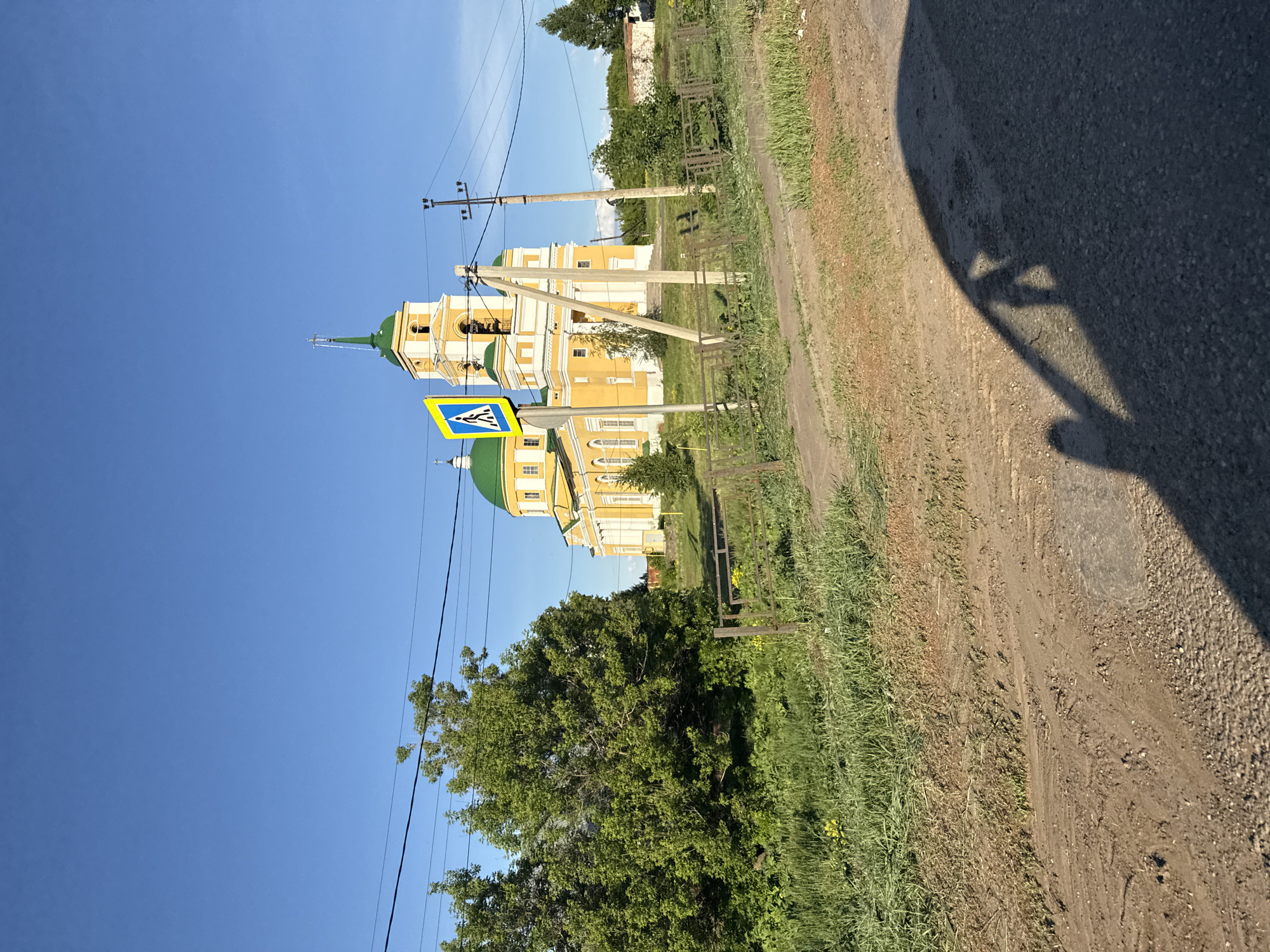 The image shows a sunny, outdoor scene in Мазунино, Russia. In the background is a yellow and white church with a green dome. A pedestrian crossing sign is next to the church. Power lines run across the sky. The foreground includes a dirt road, with green grass and vegetation on the side. A shadow of what appears to be a car is visible in the upper right corner.