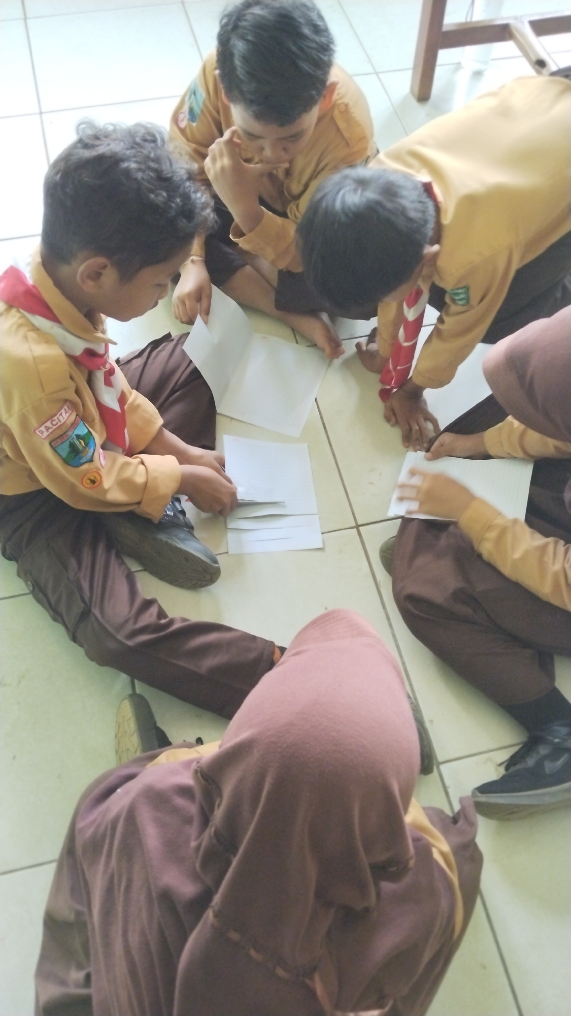 Five individuals, appearing to be young people in scout uniforms, are seated on a tiled floor, engaged in an activity with several sheets of paper. The uniforms consist of tan shirts with dark brown pants and a red and white neck scarf. One individual is leaning forward, resting their chin on their hand, with their gaze directed downwards. Another is hunched over, interacting with papers. The scene appears to be indoors, possibly a classroom or community hall, with pale floor tiles and hints of a wooden structure in the background, potentially part of a table or chair. The lighting suggests an indoor setting with overhead illumination. The setting is likely within the city of Tegalombo, Indonesia, given the context of scout activities.