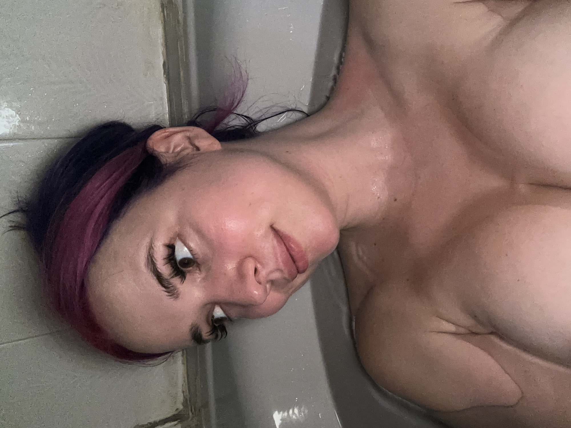 This is a close-up, low-angle, over-the-shoulder selfie of a woman in a bathtub. She has dark hair with pink streaks, and is looking to her left. Her skin is wet and appears flushed. The bathtub is white, and the wall behind her is textured and gray. The scene is likely indoors, in a bathroom.