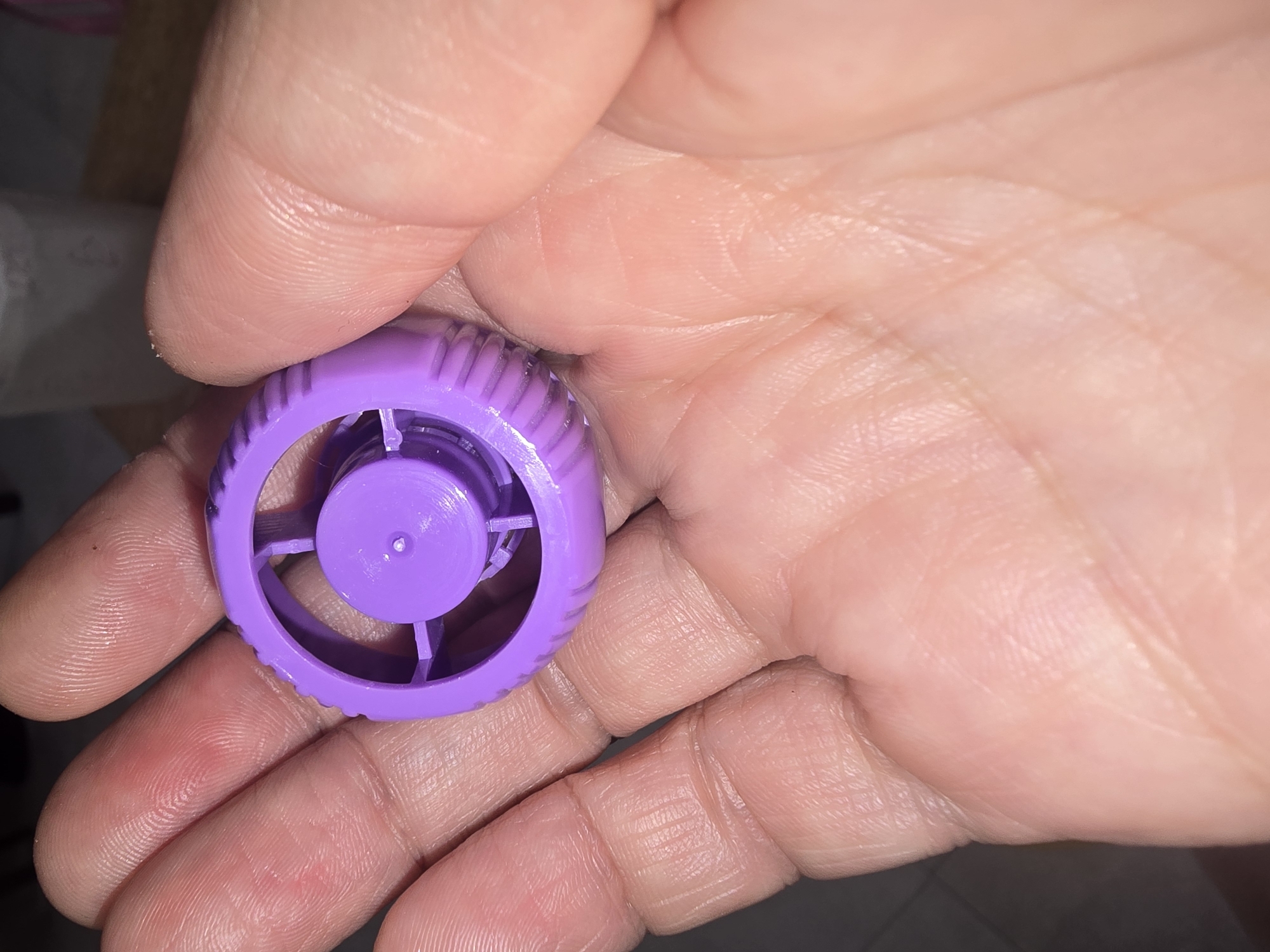 A close-up, top-down shot shows a person's hand holding a small, purple plastic cap with a ribbed outer edge and a circular, hollow center. The cap is made of plastic and has a textured surface. The hand is open, with fingers curled around the cap. The skin of the hand appears dry and wrinkled. The background is blurred and dark, with some indistinct shapes visible. There is no discernible information about the location in Ulan-Ude, Russia.