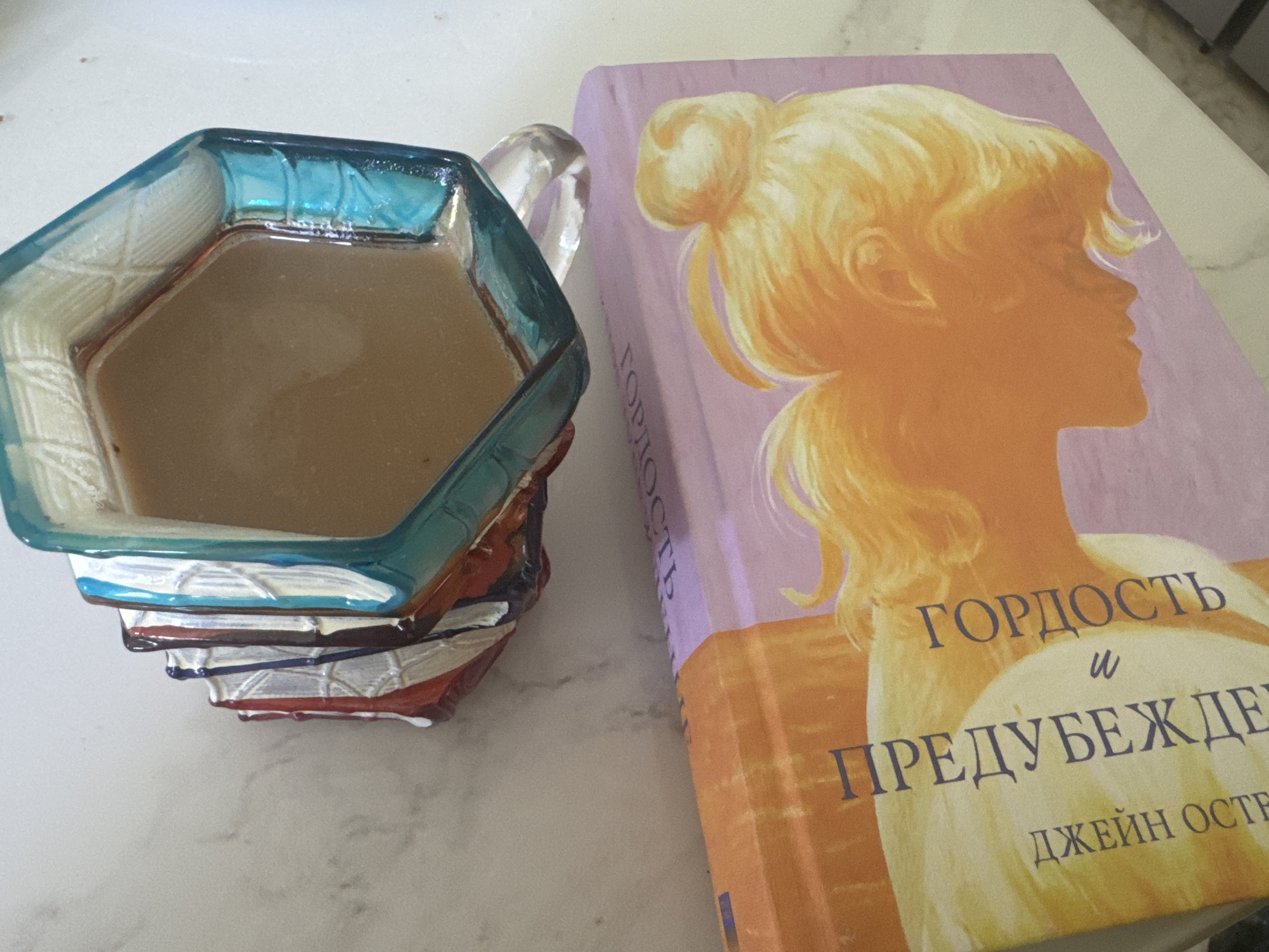 A close-up, overhead shot shows a faceted, translucent blue glass mug filled with light brown liquid, likely coffee, resting on a light-colored, marbled surface. The mug is positioned over an open book, partially obscuring it. The book has a predominantly pink cover with an illustration of a silhouette of a blonde woman with her hair in a bun. Russian text is visible on the book cover. To the left of the mug, a stack of what appears to be candy wrappers, in red, white, and blue, is stacked. The overall lighting is bright, suggesting an indoor setting with overhead illumination. There are no people or animals visible in the frame. The city of Lozhki, Russia, is not depicted in this image.