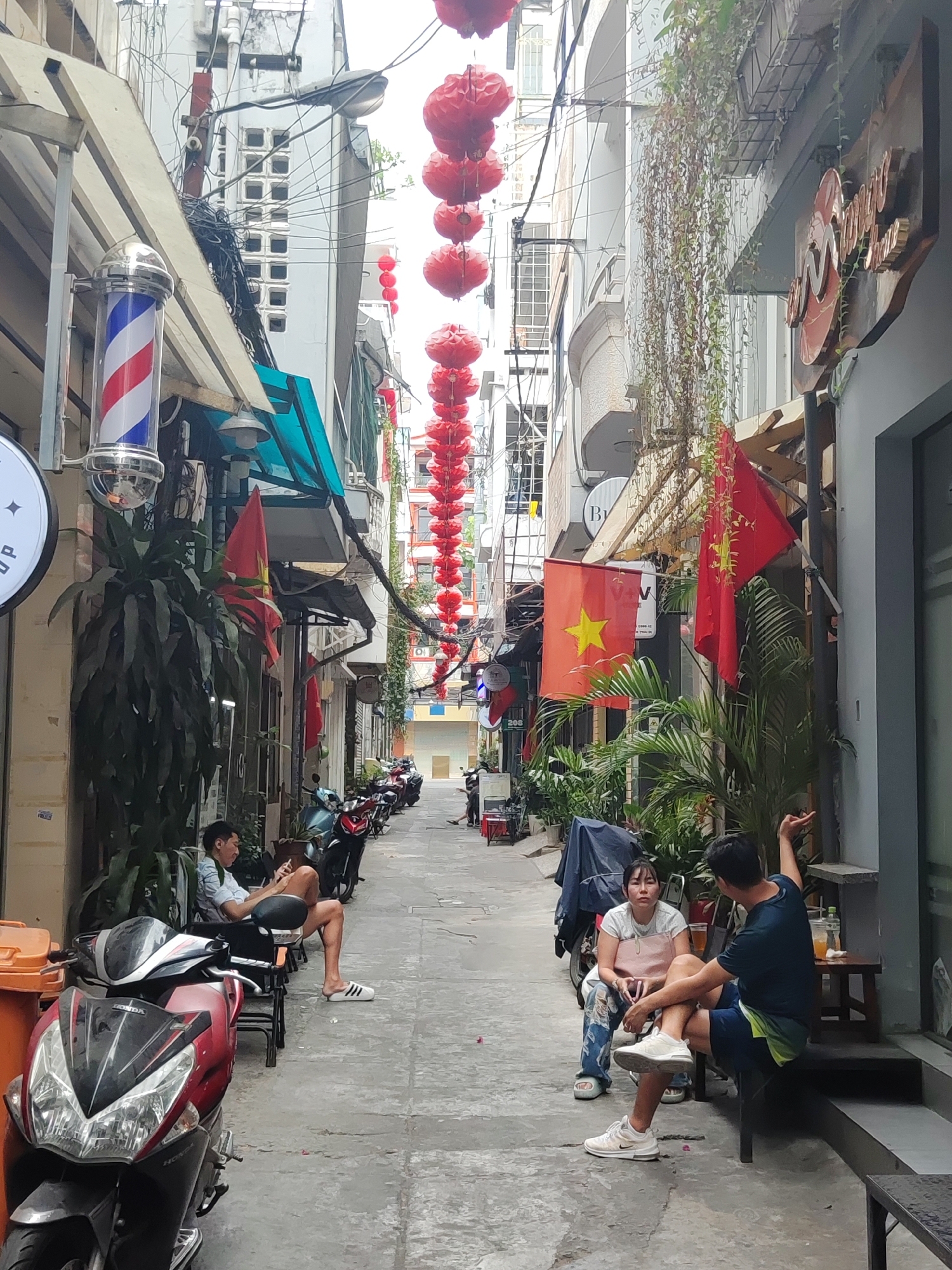 Captured in the city of Thủ Đức, Vietnam, this image depicts a narrow alleyway adorned with strings of red lanterns suspended overhead. The alley is lined with buildings of varying heights and architectural styles, some displaying Vietnamese flags. On the left, a barber pole indicates a nearby establishment. Several parked motorbikes are positioned along the alleyway's edge.
Two individuals sit on a bench on the right side of the alley, engaged in conversation. Nearby are two plastic cups containing orange juice. On the left, another person sits on a chair, looking at a phone.