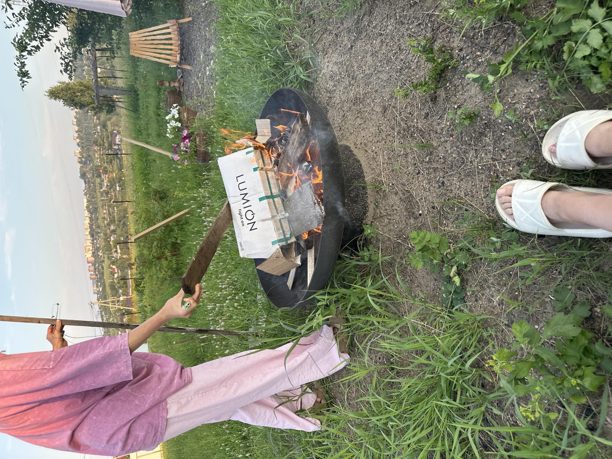 A person in a pink robe and striped pink pants is tending to a fire in a fire pit. The fire is burning a cardboard box with "LUMION light on" printed on it, along with other wooden materials. The scene is set outdoors in a grassy area with some dirt patches. In the background, there is a view of a town or city in the distance, suggesting a rural or suburban setting. The sky is visible, indicating it is daytime, likely late afternoon or early evening given the lighting. The overall atmosphere appears relaxed and informal, perhaps a casual gathering or outdoor activity. There are two pairs of feet visible in white slides in the foreground, suggesting the presence of at least one other person. The person tending the fire is holding a wooden stick, possibly to move the burning materials.