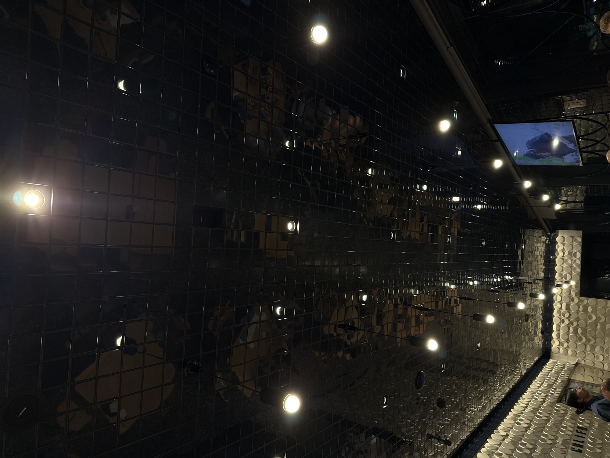 A distorted perspective of a modern interior. The ceiling is covered in dark, reflective square tiles that give off multiple reflections of lights and other objects. A row of pendant lights hangs from the ceiling, casting a warm glow. A television screen mounted near the corner of the ceiling displays a landscape scene. A textured wall with white circular patterns is located on the right side of the frame. At the bottom right, reflections of people are visible in a shiny floor, suggesting they are looking up at the ceiling.
Based on the word “California” seen on the floor, this image is likely taken inside the "California" restaurant in Querétaro, Mexico.