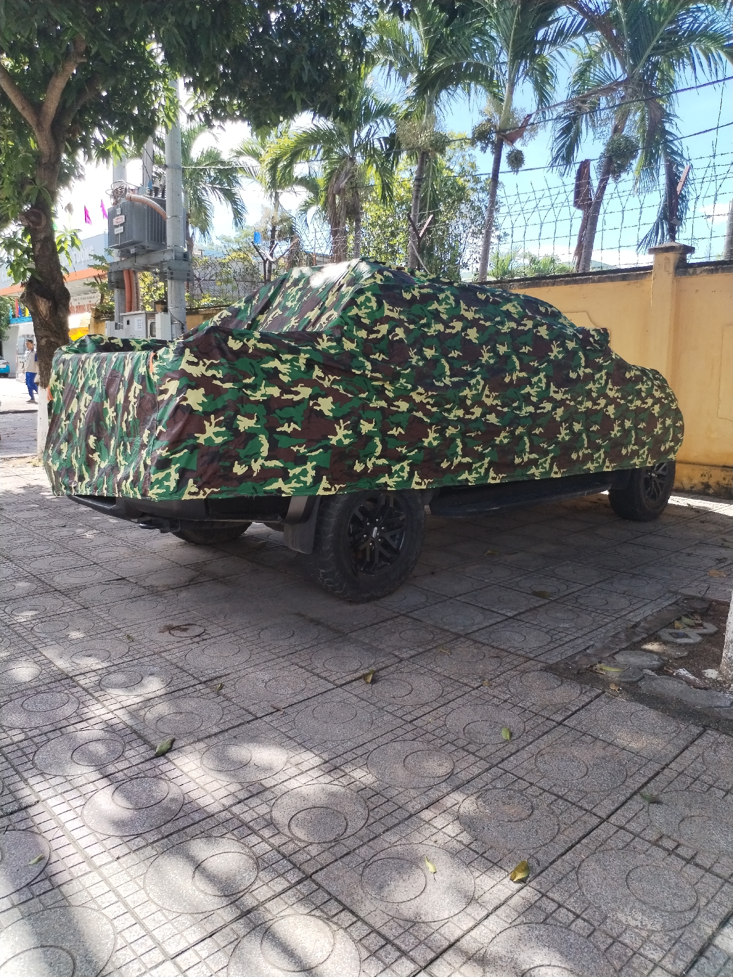 The image depicts an outdoor urban or suburban scene, likely in Nha Trang Ward, Vietnam, under bright, sunny daylight. The central subject is a vehicle, possibly a pickup truck or SUV, completely covered by a camouflage-patterned tarp in shades of green, brown, and light yellow. It is parked on a grey concrete sidewalk characterized by distinctive grid and circular patterns, with some fallen leaves scattered across it. The vehicle's dark-colored wheels are visible beneath the cover.
In the background on the left, a large, dense tree casts some shade onto the ground. Behind the tree stands a utility pole with a grey electrical transformer mounted on it. Further down the sidewalk, a person in blue attire is partially visible walking. To the right of the vehicle, a light yellow wall topped with barbed wire fencing creates a boundary. Beyond this wall, several tall palm trees reach towards a clear, bright blue sky, indicating a tropical or sub-tropical climate. The strong sunlight creates clear, defined shadows across the pavement and vehicle. The scene is tranquil, showing a typical daytime street environment. No clearly legible text is visible on any signs or objects within the frame.