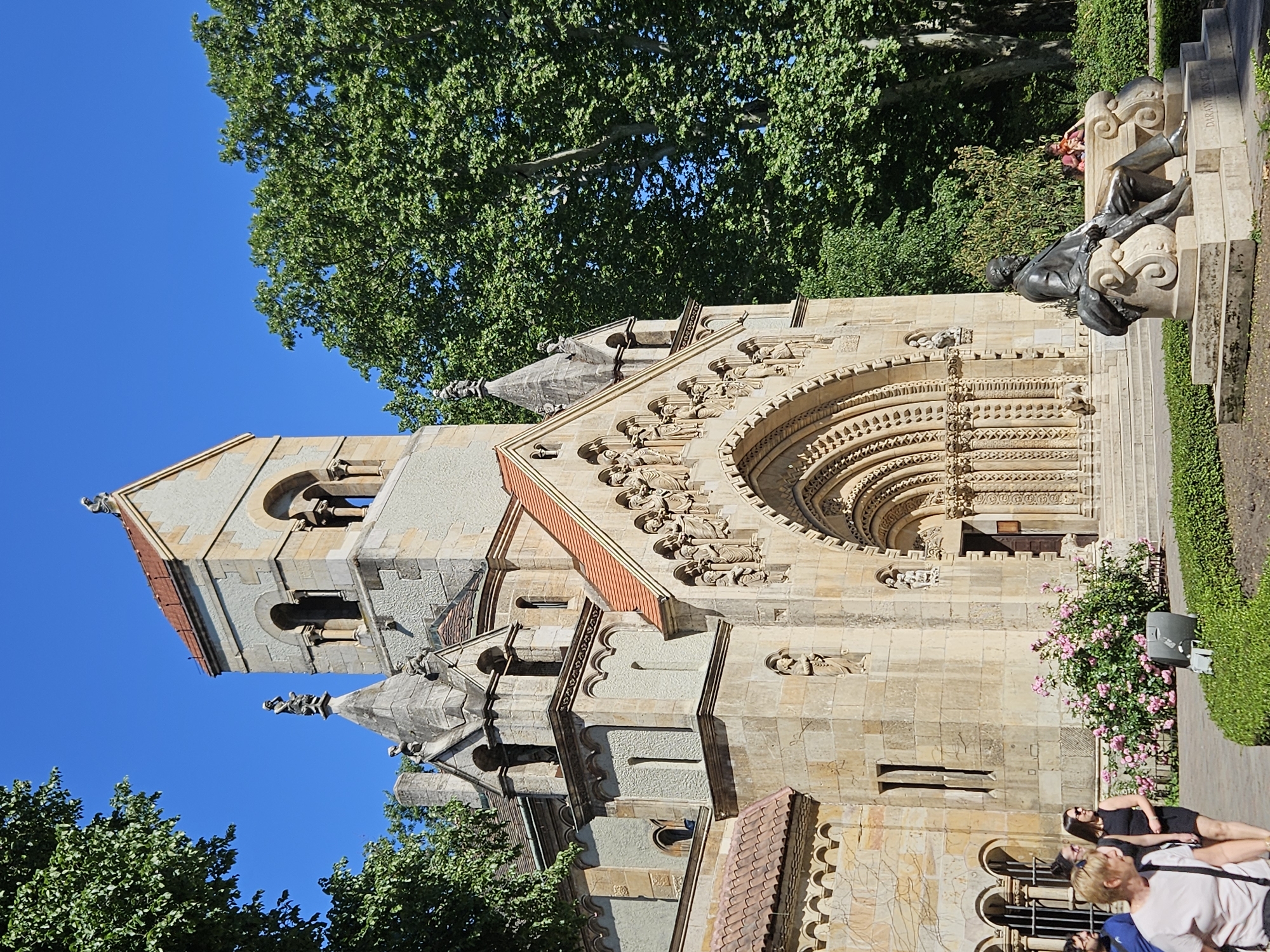 The image depicts Vajdahunyad Castle, specifically the Romanesque Chapel, located in City Park (Városliget), Budapest, Hungary. The chapel, with its detailed carvings and stone facade, is the primary architectural element. A statue of a man reclines on a raised platform to the right of the chapel. Several people are present near the bottom of the frame, seemingly observing or passing by the structure. Lush green trees frame the top and left edges of the image, contrasting with the clear blue sky. The sunlight illuminates the facade of the chapel, creating sharp shadows that define its texture and form.