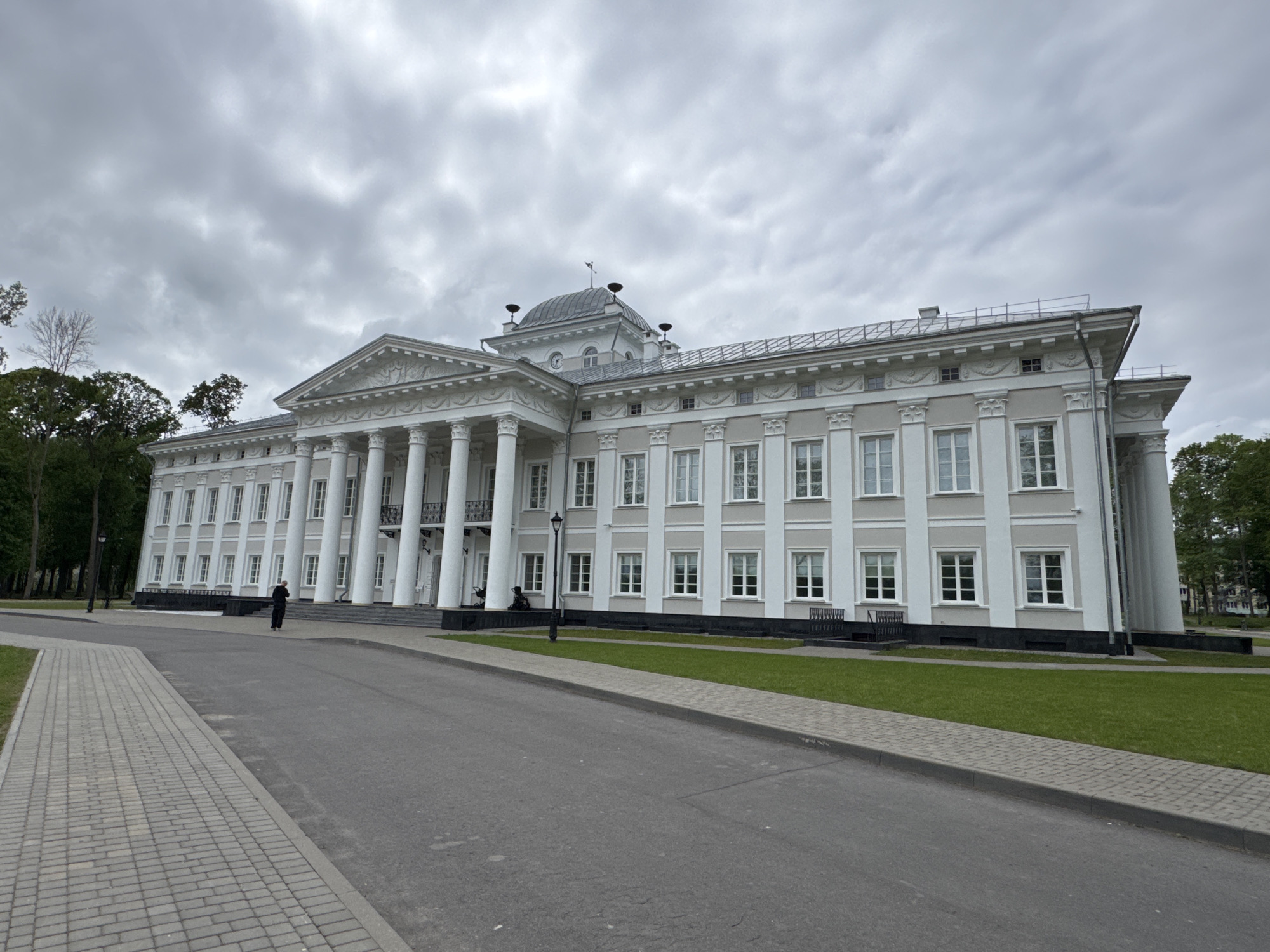 The image depicts a grand, white, neoclassical building with numerous windows and prominent columns, likely a government or historical building. The building is situated in Добасненскі сельскі Савет, Belarus. A gray paved road runs in front of it, alongside a brick-paved walkway and a patch of green lawn. The sky is overcast, casting a soft, diffused light on the scene. One person in dark clothing is walking towards the building. There are trees on either side of the building.