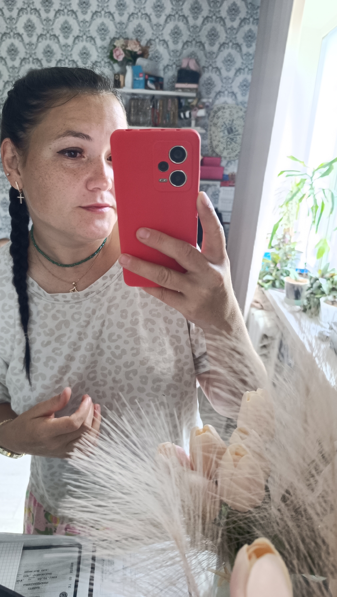 A woman is taking a mirror selfie in a room. She is holding a red smartphone in her right hand. She has dark hair in two braids and is wearing a light-colored shirt with a leopard print. She is also wearing a necklace and earrings. In the background, there is a shelf with various items, a plant, and a window. The wallpaper has a damask pattern. There is also a bouquet of flowers in the foreground. The image appears to have been taken indoors, possibly in a residential setting. There is no information in the image to suggest a specific location in the city of Chaikovsky, Russia.