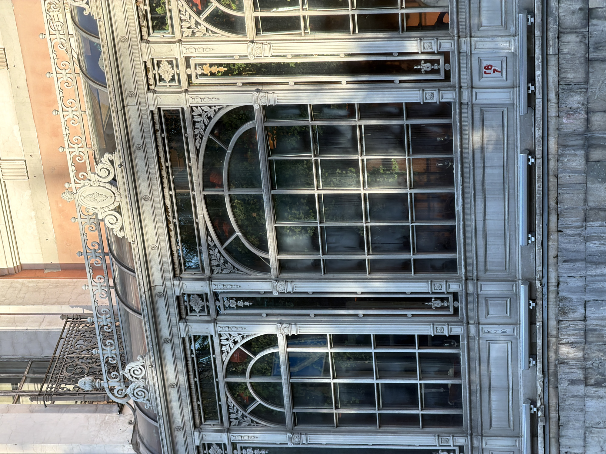 The image shows a close-up view of the ornate facade of a building, with a focus on its large, multi-paned windows and decorative metalwork. The architecture suggests a historical building, possibly from the late 19th or early 20th century, characterized by intricate detailing and a grand style. The building has a curved section, possibly a conservatory or sunroom, with a series of large windows that reflect the surroundings. These windows are divided into numerous panes by thin, dark mullions, creating a grid-like pattern. Some of the windows feature arched tops with decorative floral motifs and swirling metalwork surrounding them.
The metalwork itself is a prominent feature, appearing to be wrought iron or cast iron, painted in a muted, weathered gray. It frames the windows and forms decorative elements such as scrolling brackets and elaborate filigree along the edge of the building. Adjacent to the windows, there's a section of the building with a more classical facade, made of stone blocks. On this stone section, there are visible numbers painted in red: "18" and "7" which are likely a street address or building number. The overall impression is one of architectural grandeur and historical significance. The lighting suggests it is daytime, with sunlight casting shadows and highlighting the textures of the metal and stone. The reflections in the glass hint at greenery from an interior space, possibly a botanical garden or a well-maintained courtyard. The context provided places this scene in Moscow, Russia.