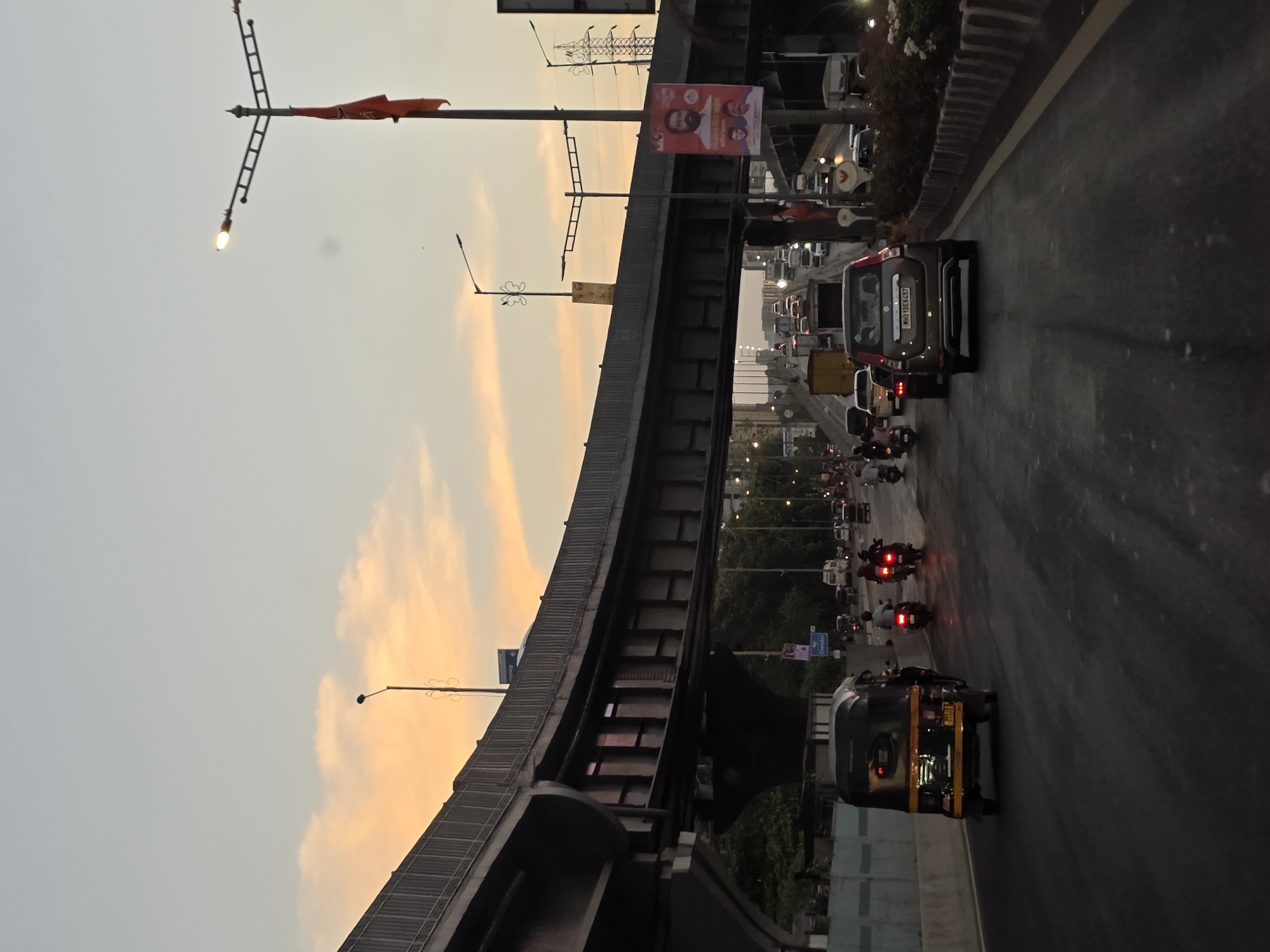 This is a vertical, eye-level, outdoor shot of a busy multi-lane road in Mumbai, India, during dusk. The sky displays a soft gradient of pale blue and orange clouds. An elevated highway with a grey concrete structure dominates the left and central portions of the frame, casting a shadow over the lower part of the road.
On the road, traffic is moderate, consisting of cars, auto-rickshaws, and motorcycles. In the foreground, a yellow auto-rickshaw with a black roof is visible, moving away from the camera. To its right, a silver SUV with a license plate that reads "MH01DE8487" is also traveling away. Several motorcycles and scooters are interspersed with the cars, some with visible riders. On the right side of the road, a decorative concrete barrier lines the edge, with some greenery and flowers visible behind it.
Streetlights are present, including a prominent one with a T-shaped fixture and a red flag attached to its pole on the right side of the image, illuminated by the fading light. A large poster featuring a portrait of a man in a white shirt and two smaller portraits below it is attached to the same pole. Other overhead power lines and poles are also visible in the distance, as well as a large electrical pylon on the far right. The overall scene depicts a typical urban commute in Mumbai as the day transitions to evening.