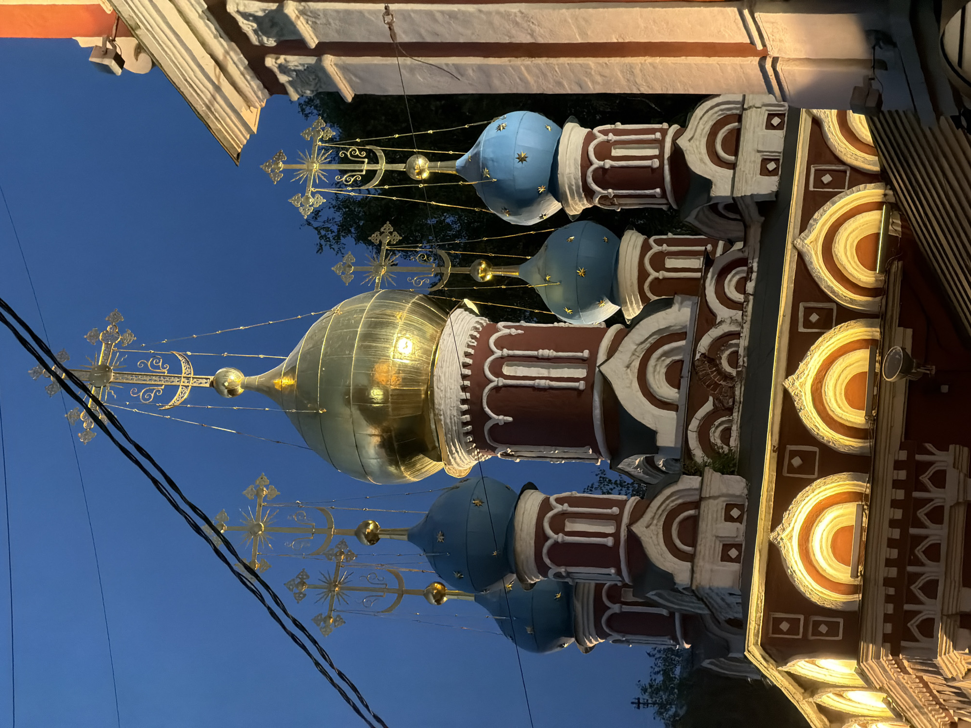 The image shows a close-up, low-angle view of the onion domes of a Russian Orthodox church at dusk or night. The sky is a deep blue, with power lines crisscrossing the upper portion of the frame. The domes are arranged in a cluster, with one large golden dome prominently featured in the center, reflecting the light. Surrounding it are several smaller, sky-blue domes topped with golden crosses. The church's facade is visible at the bottom of the frame, featuring red and white ornate decorations, including arched windows illuminated from within, and decorative cornices. The overall scene suggests an exterior shot of an active religious building, likely in Moscow, Russia, given the architectural style typical of Russian Orthodox churches. There are no people or specific actions depicted; the focus is entirely on the architecture and atmosphere.