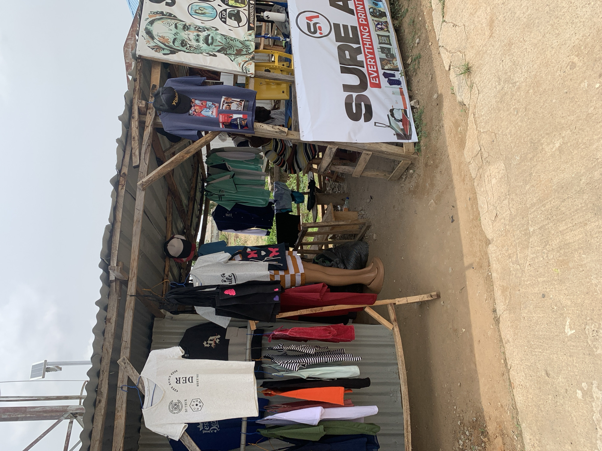 The image shows a roadside stand selling clothes and printing services in Keffi, Nigeria. The stand is constructed of wood and corrugated metal, providing shelter for the merchandise. A variety of clothing items, including t-shirts, shirts, and dresses, are displayed on hangers and on a mannequin torso. Banners advertise "SURE A EVERYTHING PRINT" and show examples of printed products like hats and portraits. The ground is a mix of dirt and pavement. A solar-powered light fixture is visible near the top of the frame.