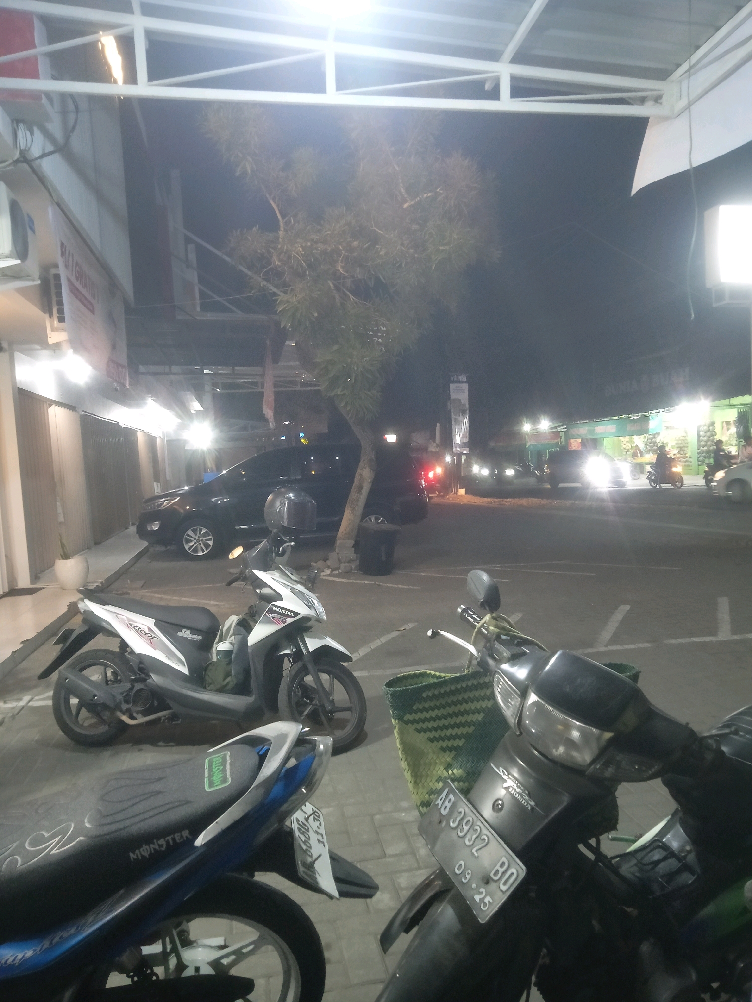 The image depicts a nighttime scene in front of a row of buildings, possibly shops, on a street in Trihanggo, Indonesia. The foreground features two parked motorcycles. The closer motorcycle is black with a green woven basket attached to the handlebars. Its license plate reads "AB 3932 BO 09-25". The second motorcycle, slightly behind and to the left, is white and silver. A black car is parked further back in the scene, and beyond that, the street is visible with other vehicles, including motorcycles and cars, some with headlights illuminated. One building has bright lighting and displays signs, one of which appears to read "DUNIA BUAH" (Fruit World). A tree with sparse foliage stands between the parked cars and the street. The overall environment suggests a commercial area, possibly a market or shopping strip.