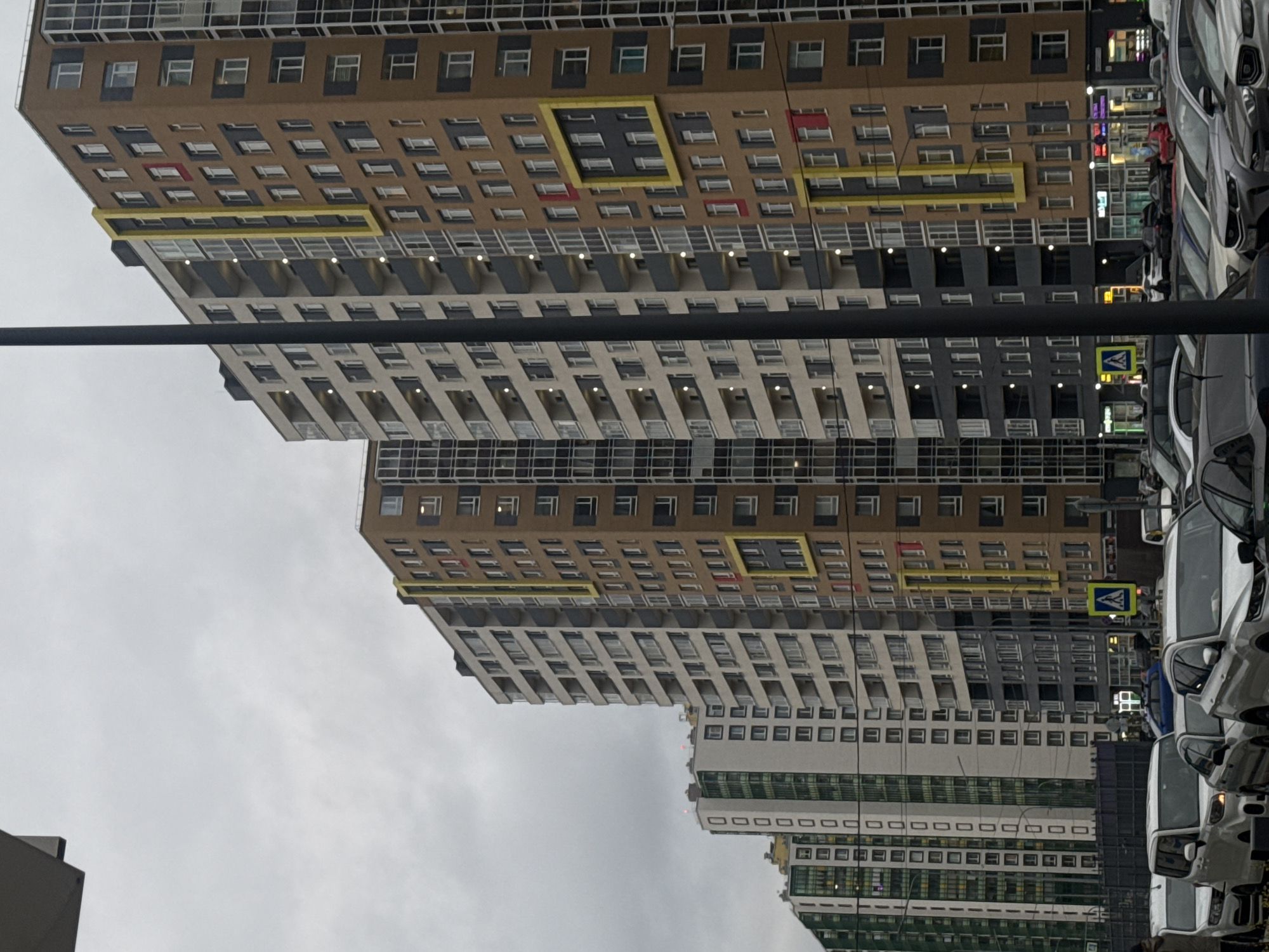 This image captures an urban street scene in Murino, Russia, characterized by modern, high-rise residential buildings and a street lined with parked cars, all under an overcast sky.
The dominant features are several multi-story apartment buildings. The most prominent building, occupying much of the left side of the frame, has a complex facade. Its upper sections are brown with numerous rectangular windows, some of which are highlighted with bright yellow and red framing. Below this, the building transitions to lighter shades of grey and white, featuring multiple rows of balconies equipped with small, illuminated lights, suggesting either dusk or a very dim daytime. Further into the background, other similar tall buildings are visible, some incorporating green-striped patterns on their lower facades.
To the right, a row of cars is parked tightly along what appears to be the roadside or a parking lot. The vehicles are mostly light-colored, predominantly white and grey.
A thick, dark horizontal bar, possibly a utility pole or overhead structure, traverses the mid-ground of the image. Below this bar, near the street level, a yellow square sign with a black border and a black pedestrian icon is visible, indicating a pedestrian crossing. The sky is uniformly grey and overcast, suggesting a cool or non-sunny day. The overall scene depicts a densely populated, modern residential area.