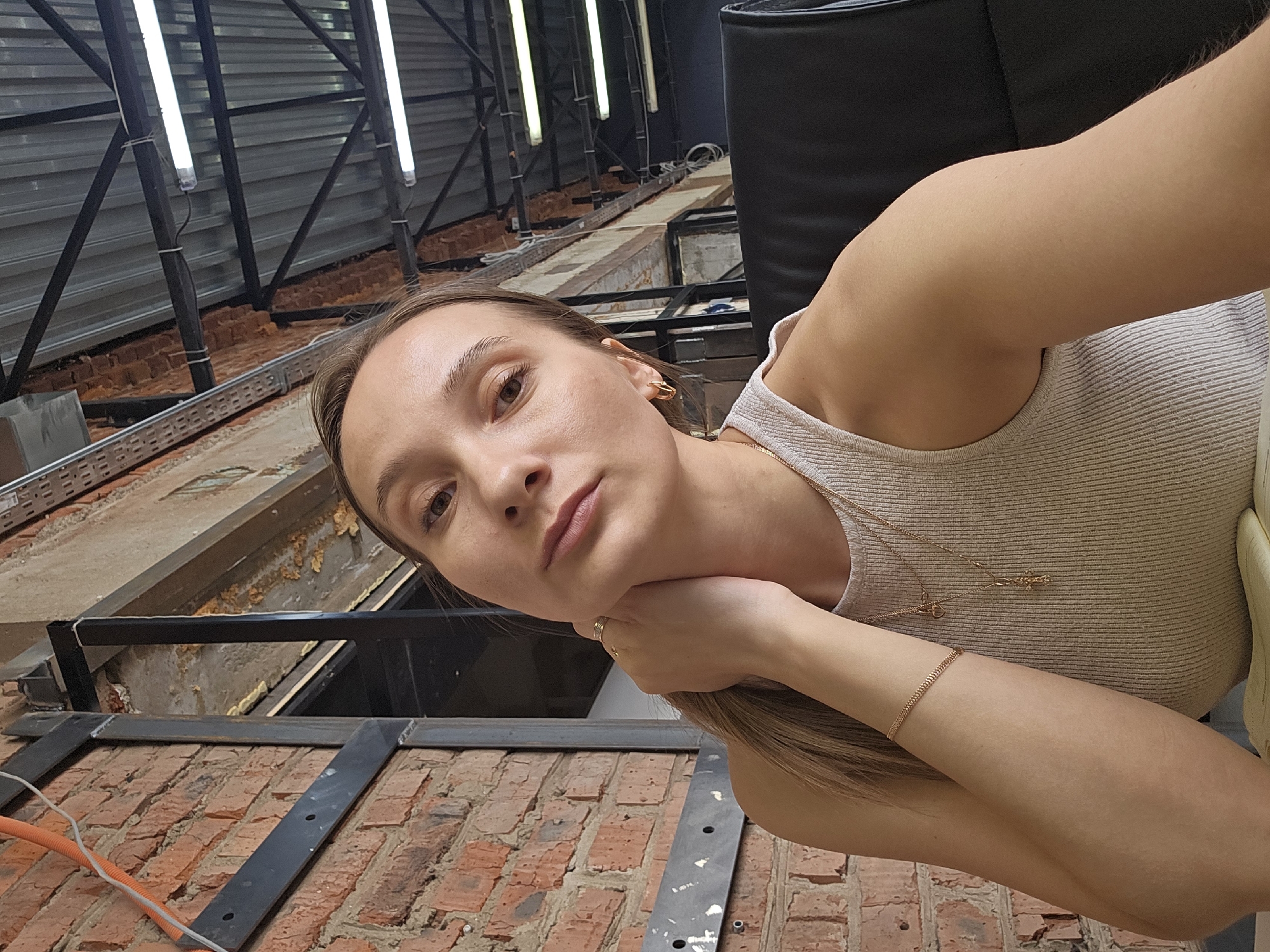 This image depicts a young adult female, with light skin and light brown hair, posing indoors in Chaikovsky, Russia. She wears a light-colored ribbed tank top and is adorned with gold hoop earrings, a layered gold chain necklace featuring a cross and a heart pendant, a gold bracelet on her right wrist, and a gold ring on her left ring finger. Her left hand is positioned gently against the right side of her neck, and she looks directly into the camera. The background reveals an industrial-style interior: an exposed red brick wall on the left is partially covered by black metal structural elements and an orange electrical conduit. Overhead, a corrugated metal ceiling with black trusses and multiple fluorescent tube lights is visible. A black office chair back is present behind her right shoulder, and a section of a light-colored notebook is visible at the bottom edge of the frame.