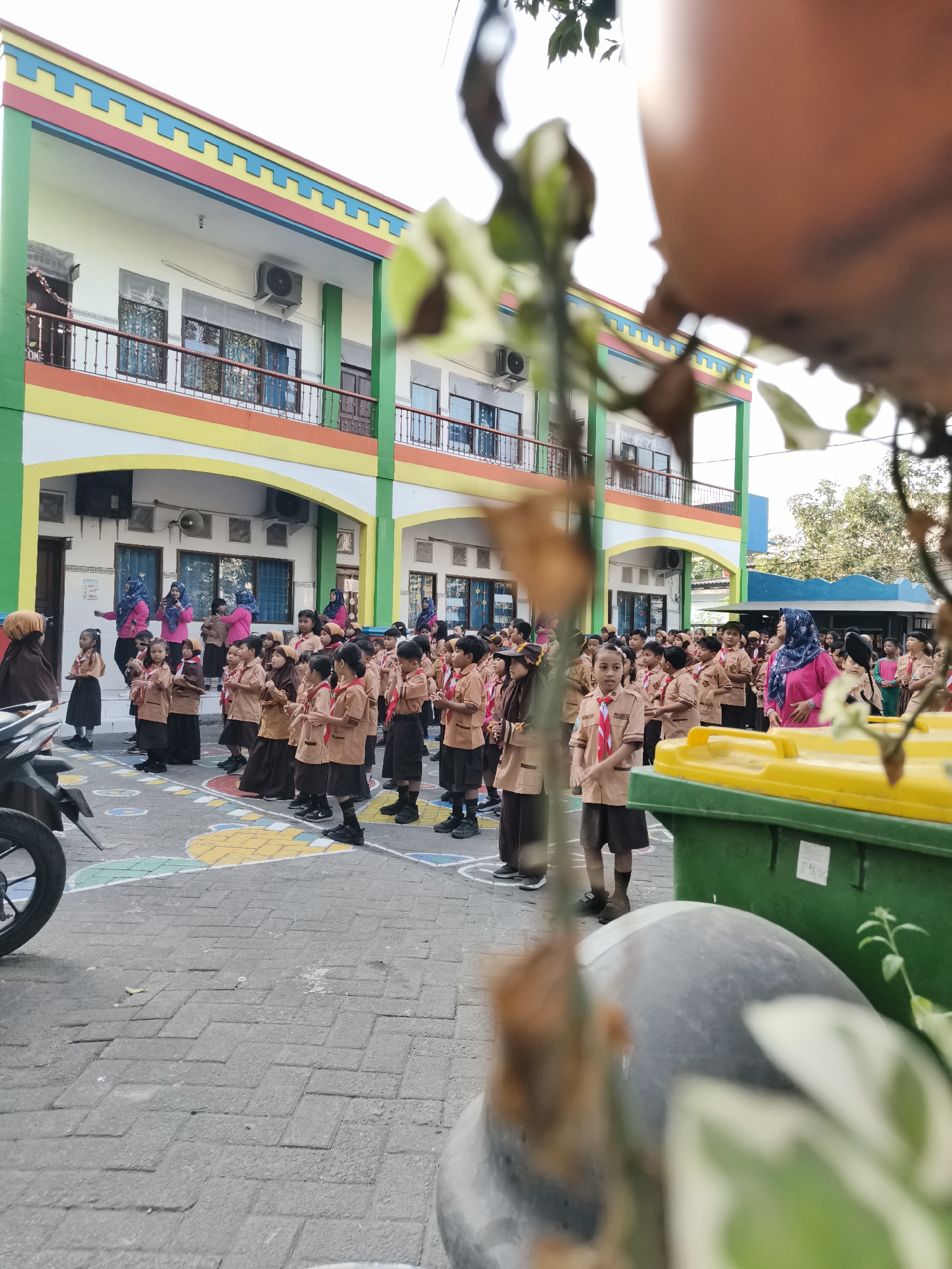 In Petiken, Indonesia, a group of young students in uniform stand in formation on an outdoor paved area. The students, predominantly girls, wear light brown shirts and dark skirts or shorts, some with red and white neckerchiefs, indicating they are likely part of a scouting or similar youth organization. They are arranged in multiple rows, facing a building in the background.
The building appears to be a school, painted in bright colors with a multi-story structure featuring windows and balconies. A colorful decorative border, incorporating yellow, red, and blue, runs along the top of the building's facade. In front of the building, several adults, presumably teachers or supervisors, are visible. Some are wearing pink shirts and headscarves, and at least one appears to be giving instructions.
To the right of the students, two large green trash bins with yellow lids are positioned. A motorcycle is parked on the left side of the frame, and the foreground is partially obscured by the out-of-focus leaves and a pot, suggesting the photo was taken from behind some foliage. The ground is paved with interlocking bricks, and colorful painted shapes are visible on the ground in front of the students. The overall scene depicts an outdoor assembly or activity for school children.