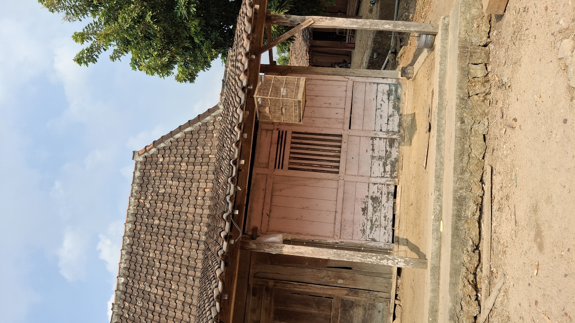 The image shows a traditional Indonesian house in Pucakwangi, Indonesia. The house has a red tile roof and is made of wood, with a weathered pink facade. There is a birdcage hanging from the porch. The house is situated on a dirt ground, with a concrete step in front of it. In the background, there is a lush green tree and another similar house. The sky is clear with some scattered clouds. The scene appears to be a rural or semi-rural setting.