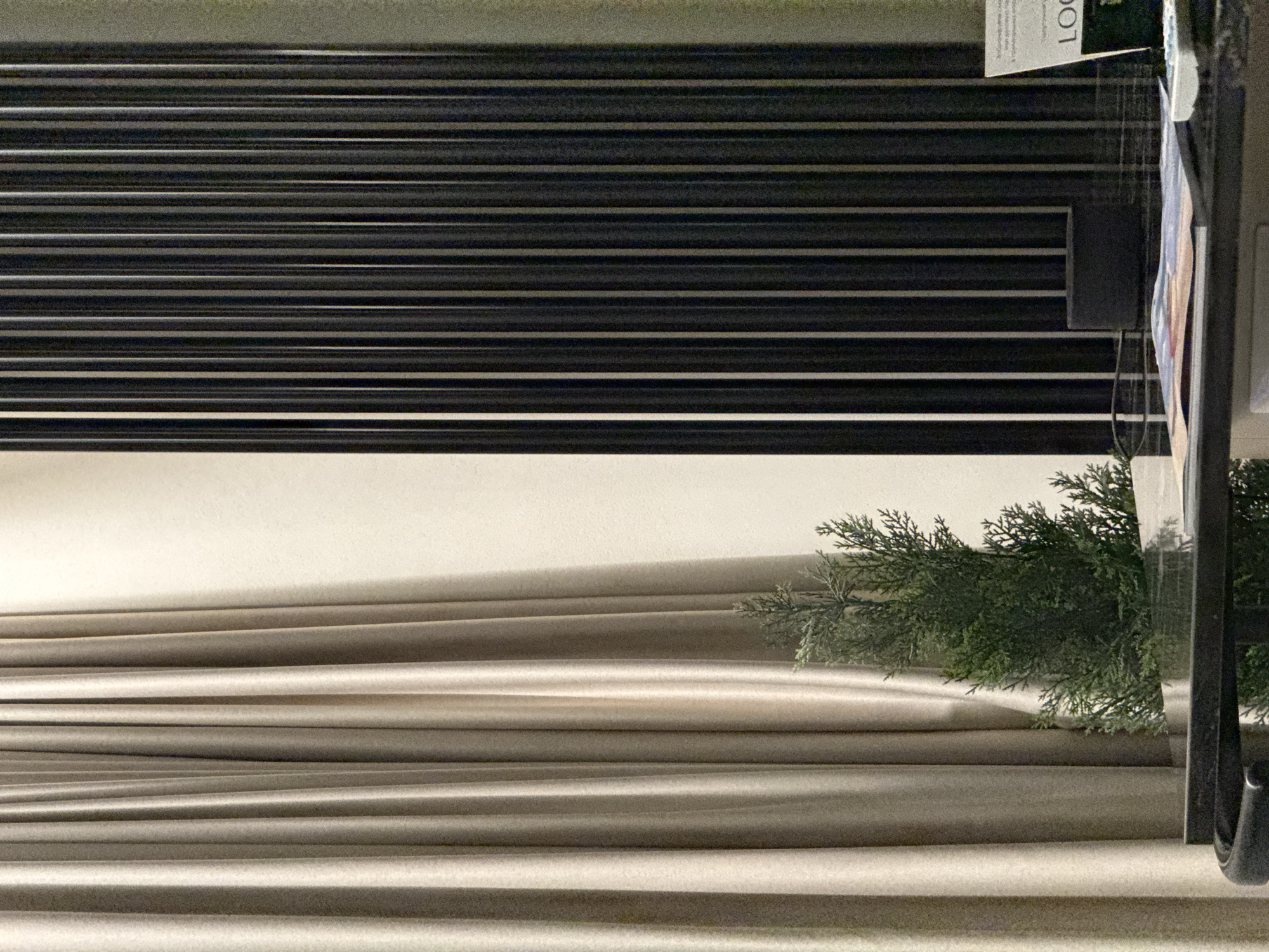 The image depicts a close-up view of a modern interior, possibly a hotel room or a reception area. On the left, a light beige curtain is draped, creating soft folds. To the right of the curtain, a vertical slatted partition in a dark color, likely black or dark gray, stands prominently. A small, artificial evergreen tree is positioned in front of the curtain on a dark, reflective surface, likely a table or desk. On the table, there are some papers or magazines, a small dark cylinder, and a white object with buttons, possibly a calculator or a remote control. To the far right, a white sign with dark text is visible, with the word "LOG" clearly legible. The lighting appears to be subdued, casting soft shadows. There are no people visible in the image. The scene suggests a sophisticated and minimalist aesthetic. The image has been taken indoors and provides no direct visual cues to pinpoint its specific location within the city of Izhevsk, Russia.