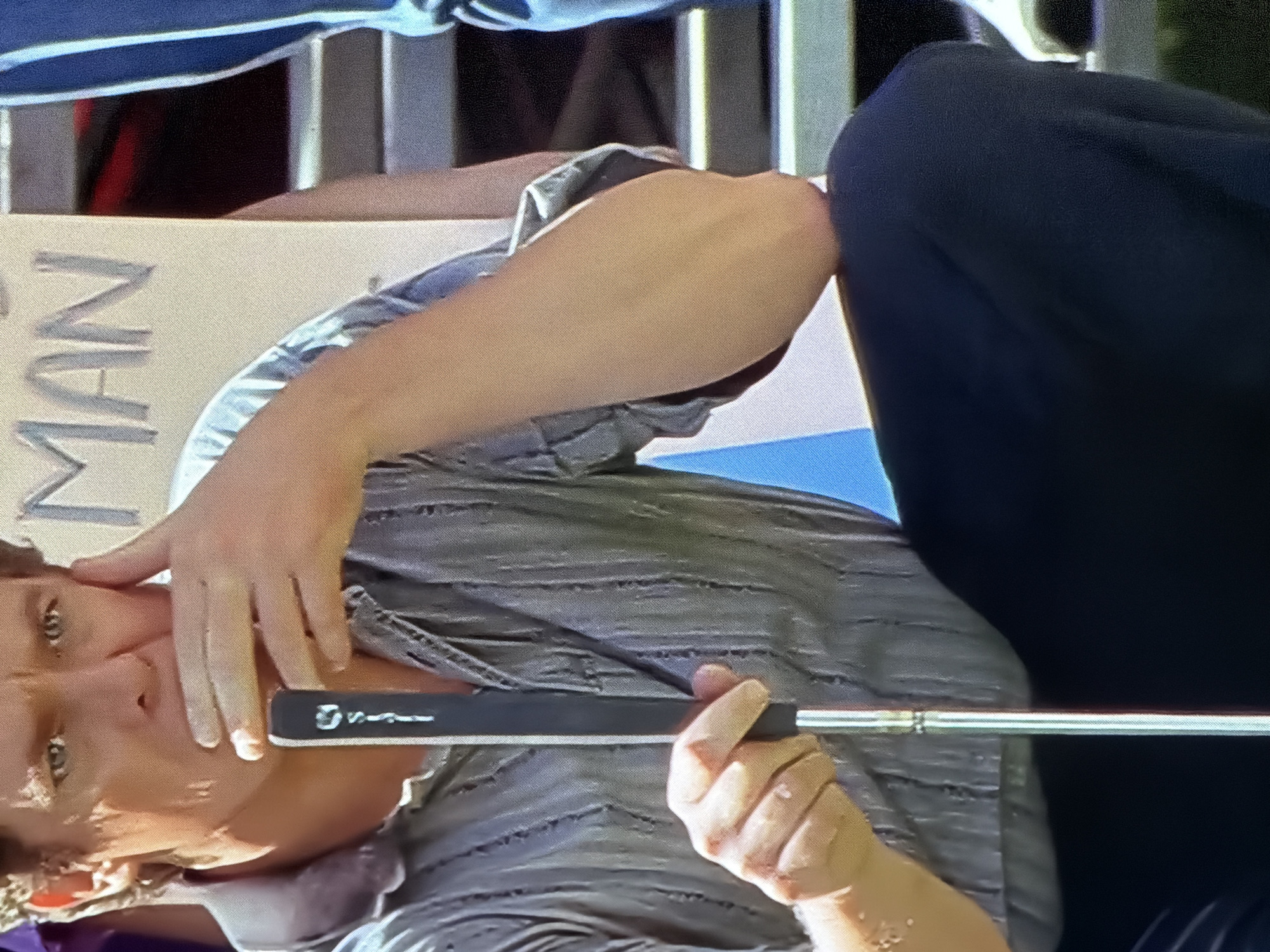 A man is kneeling, holding a golf putter. He is wearing a gray striped shirt and dark pants. His left hand is on the putter, and his right hand is touching his face. In the background, a sign reads "MAN". The scene appears to be outdoors, possibly on a golf course or a public event. The location in the city of Chaikovsky, Russia, is not ascertainable from the image.