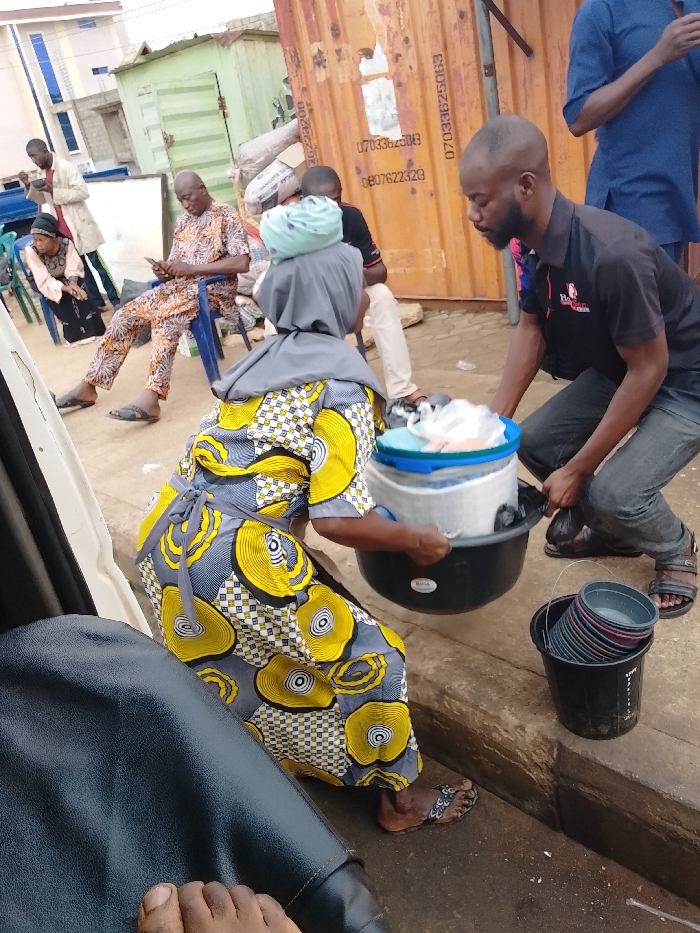 A woman in a yellow patterned dress and gray head covering is assisted by a man in a black shirt, as they carry a large black bowl containing a white bucket with a blue lid. Another bucket filled with bowls sits on the ground. Several people are sitting and standing in the background, with buildings and a cargo container visible. The picture appears to be taken from inside a vehicle, as a leather car seat and feet are partially visible in the foreground.