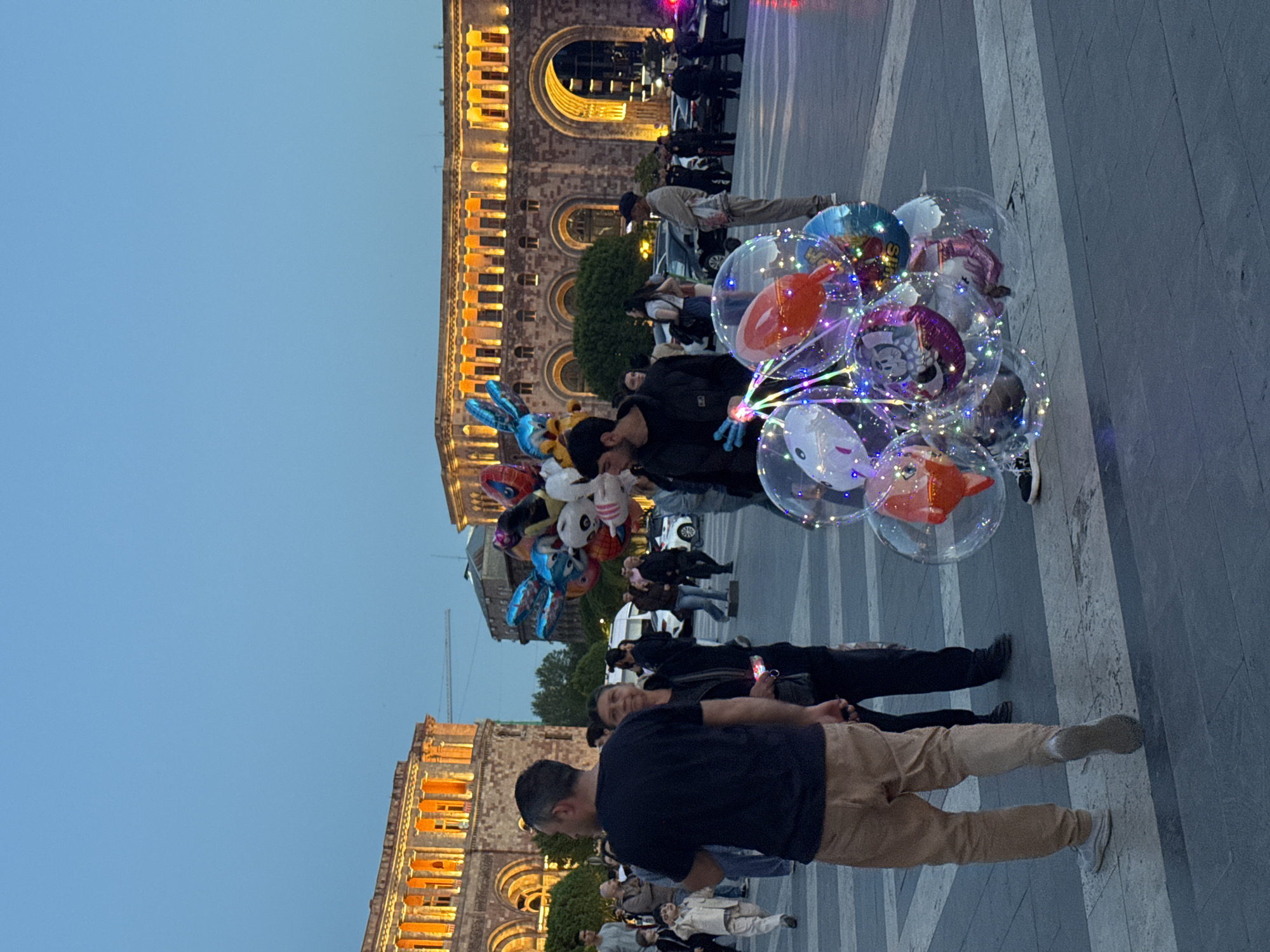 The image shows a group of people in Republic Square, Yerevan, Armenia. Several people are standing and walking. A man is holding a bunch of decorative balloons, including clear ones with internal lights and others with character designs. The State Revenue Committee building is visible in the background. The sky is light blue.