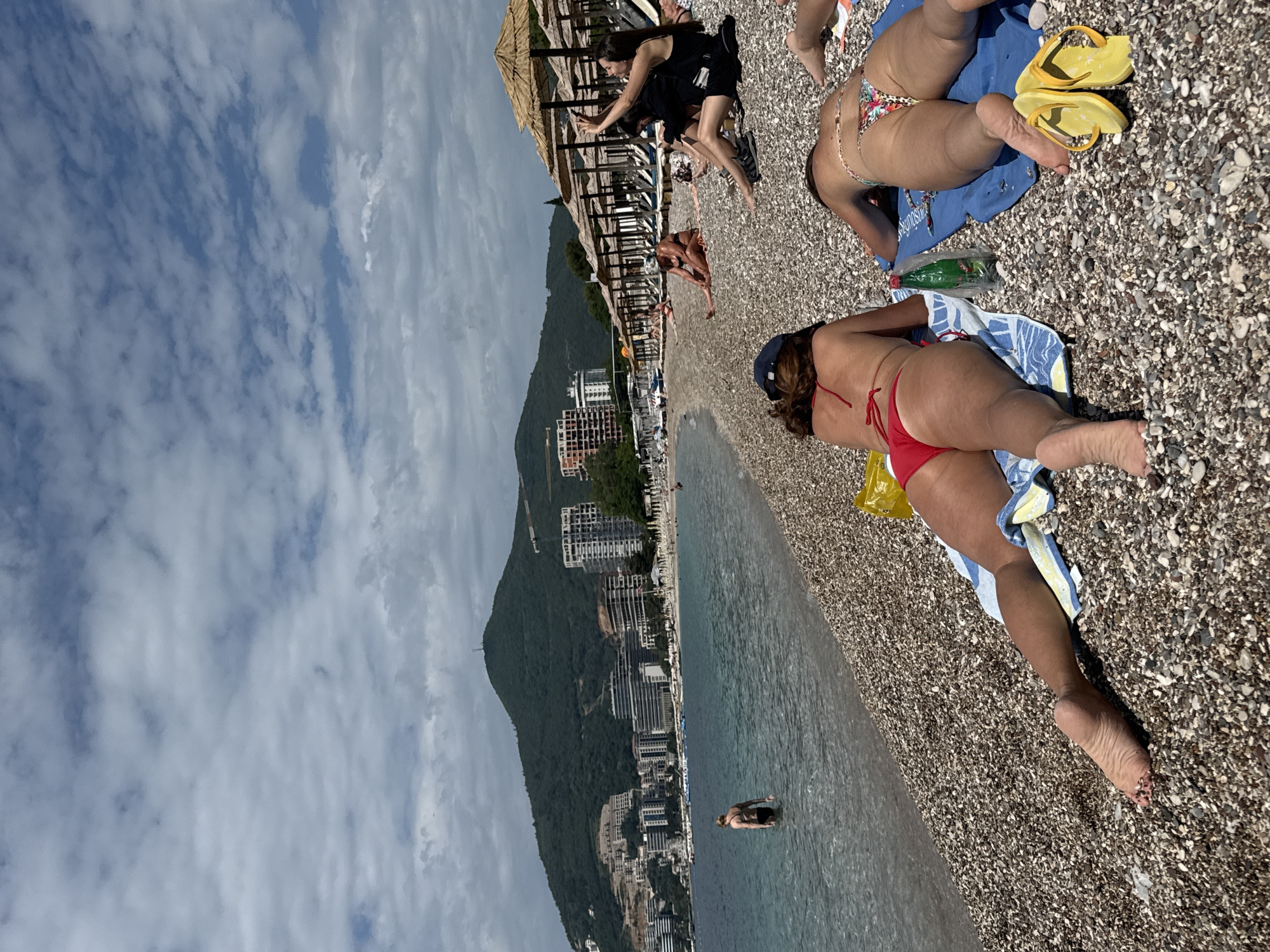 The scene captures a sunny beach in Budva, Montenegro, characterized by a pebble shoreline and the Adriatic Sea meeting the coast. In the foreground, several individuals are relaxing on the beach; one woman is lying on a towel wearing a red bikini, while another is also reclining on a towel in a floral bikini with yellow flip-flops nearby. People are also seen walking and interacting near beach umbrellas with thatched roofs. Buildings and lush mountains stretch along the coastline, contributing to the landscape. Clouds lightly pepper the blue sky overhead. Further in the water is another swimmer.