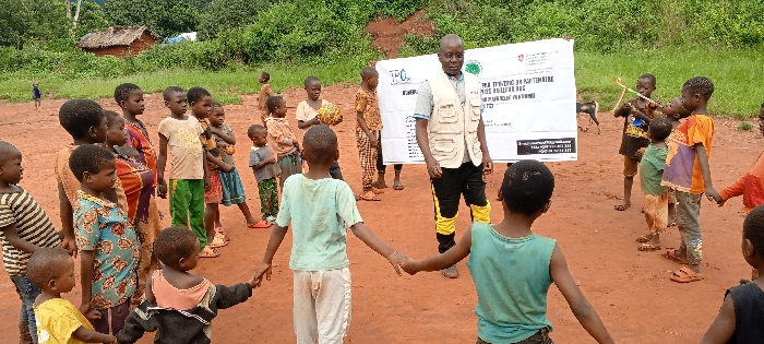 The image portrays an outdoor gathering in Ngalula, Democratic Republic of the Congo, set on reddish-brown dirt with dense green vegetation and a visible thatched-roof hut in the background. Approximately 25-30 individuals are present, predominantly children ranging from toddlers to early teenagers, and one adult male. The adult male, positioned slightly right of center, wears a light-colored shirt, black pants with yellow lower leg panels, and a multi-pocketed beige vest. He stands in front of a large white banner featuring the "TPO" logo, a Red Cross logo, another green logo, and French text, including "PROJET DE L'UNITE EN PARTENAIRE" and "DRC." A central group of 10-12 children forms a circle, holding hands, with additional children observing from various positions. One child on the far right holds a makeshift bow and arrow. A small black and white dog is visible in the background near the banner. The scene depicts an organized group activity, likely educational or recreational, for the children.