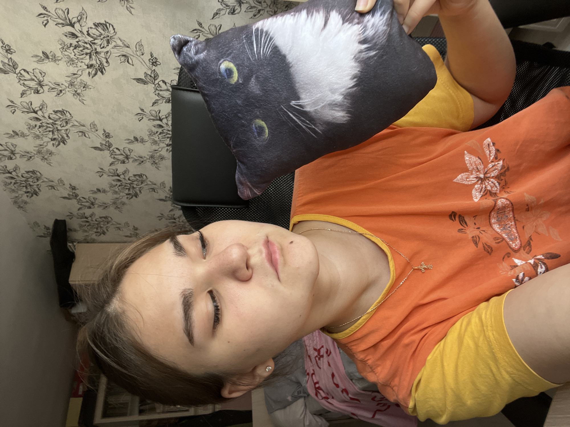 The image depicts a young woman seated indoors, likely in the city of Chaikovsky, Russia. She is positioned slightly to the left of the frame, looking downwards and to her right with a neutral expression. Her light brown hair is pulled back, revealing a fair complexion with visible freckles or moles on her face, including one prominent mole below her left lip. She has well-defined eyebrows and eyelashes. A small, clear stud earring is visible in her left ear. She is wearing a two-toned t-shirt: orange on the body with a floral graphic print in lighter tones, and yellow short sleeves and collar trim. A thin gold chain necklace with a small gold cross pendant hangs around her neck.
She holds a rectangular plush pillow, printed with the image of a black and white tuxedo cat, in her left hand. The cat on the pillow has prominent green eyes and white whiskers. The pillow is held upright, tilted slightly towards the viewer. The woman appears to be seated in a black office chair with a mesh backrest, part of which is visible behind her.
The background features a wall covered in light-colored wallpaper with a repeating pattern of dark, stylized floral or botanical motifs. To the upper left, a portion of a light-colored shelving unit or cabinet is visible, containing indistinct objects. Below this, a pink or red patterned blanket or fabric is partially visible, suggesting a bed or other soft furnishing. The scene is illuminated by ambient indoor light.